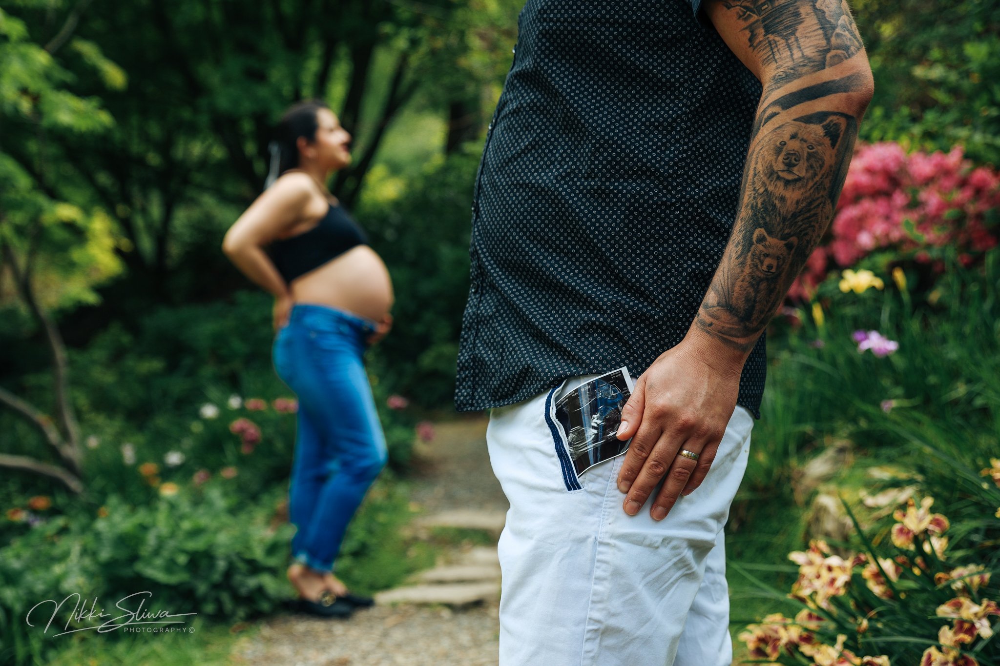 A man with tattoos on his arm holding a sonogram picture in his pocket while standing in a lush garden, with a pregnant woman in the background.
