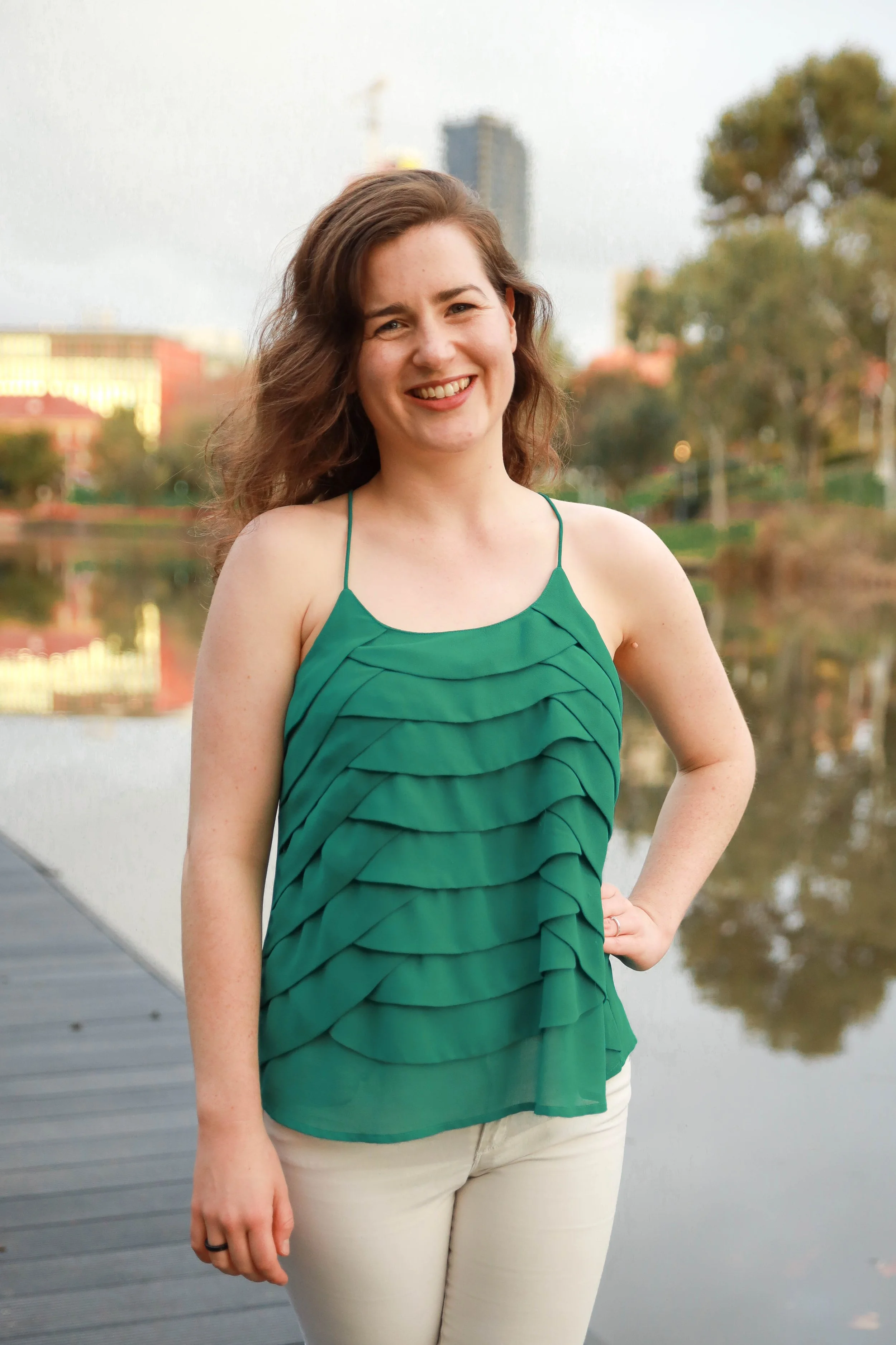 A young woman with curly brown hair smiling at the camera, wearing a green ruffled sleeveless top, standing outdoors near a body of water with trees and buildings in the background.