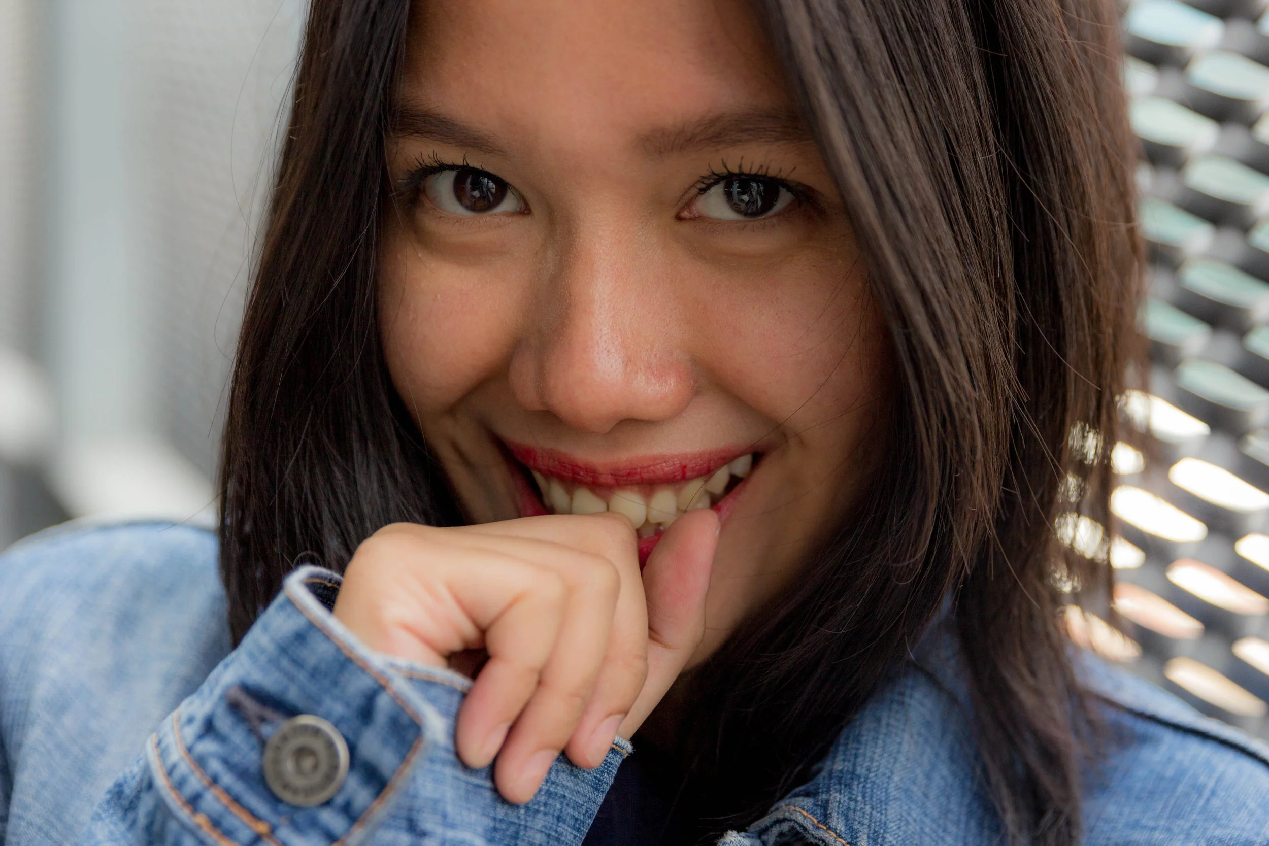 Close-up of a smiling woman with dark hair, wearing a denim jacket, holding her hand near her mouth.