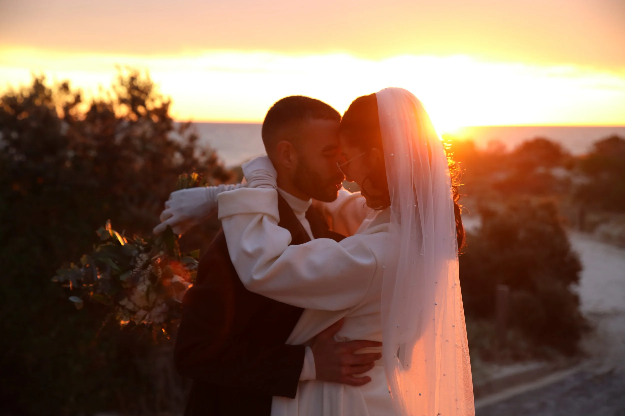A couple on their wedding day embracing each other at sunset, with the ocean in the background and warm, orange lighting.