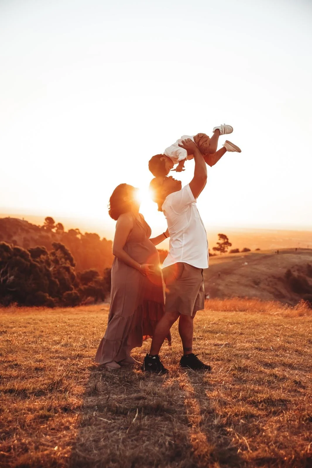 A family of three enjoying a sunset outdoors, with the father lifting a child into the air, the mother pregnant, standing on a grassy field with rolling hills and trees in the background.