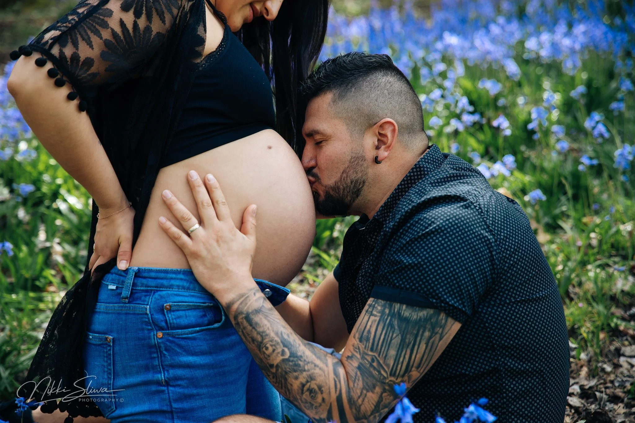 A man kisses a pregnant woman's belly while she stands outdoors in a field of blue flowers, with her lower body visible. The man has tattoos on his arm and a beard, and the woman is wearing a black top with lace sleeves and blue jeans.
