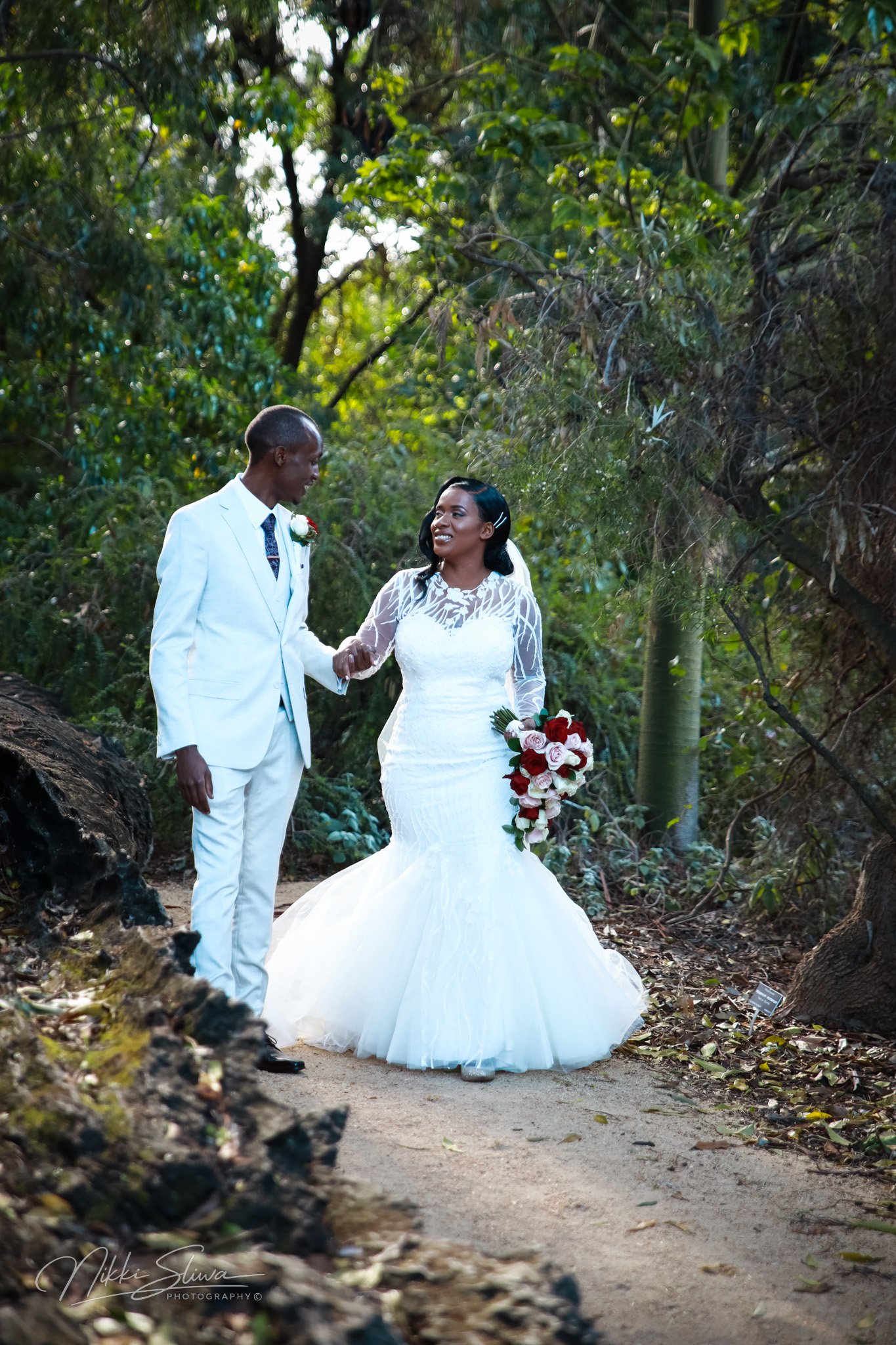 A bride and groom holding hands in a wooded outdoor setting, with the bride wearing a white wedding gown and holding a bouquet of red and white roses, and the groom dressed in a white suit.