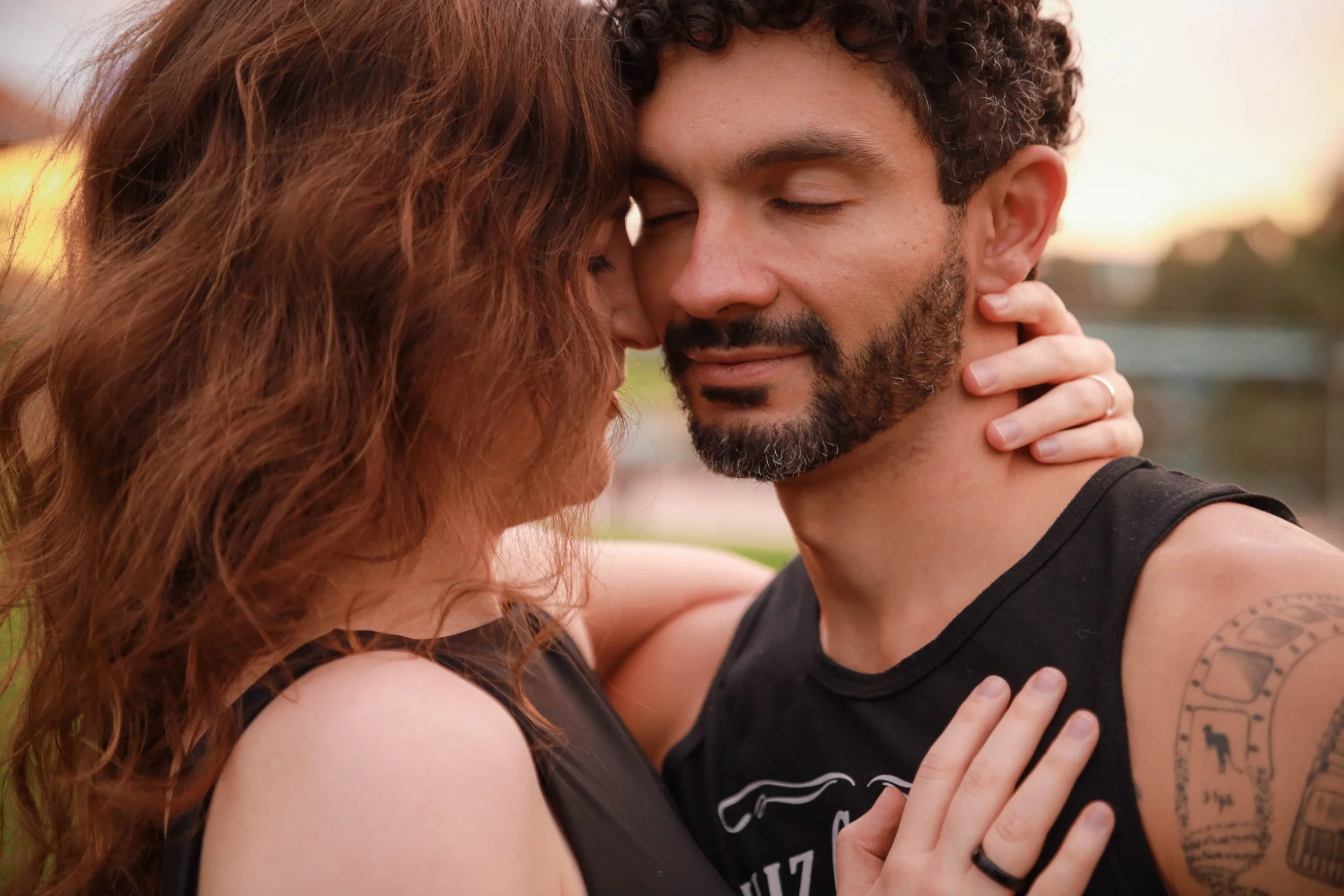 A couple embracing outdoors at sunset, with their foreheads touching and eyes closed, showing affection and closeness.