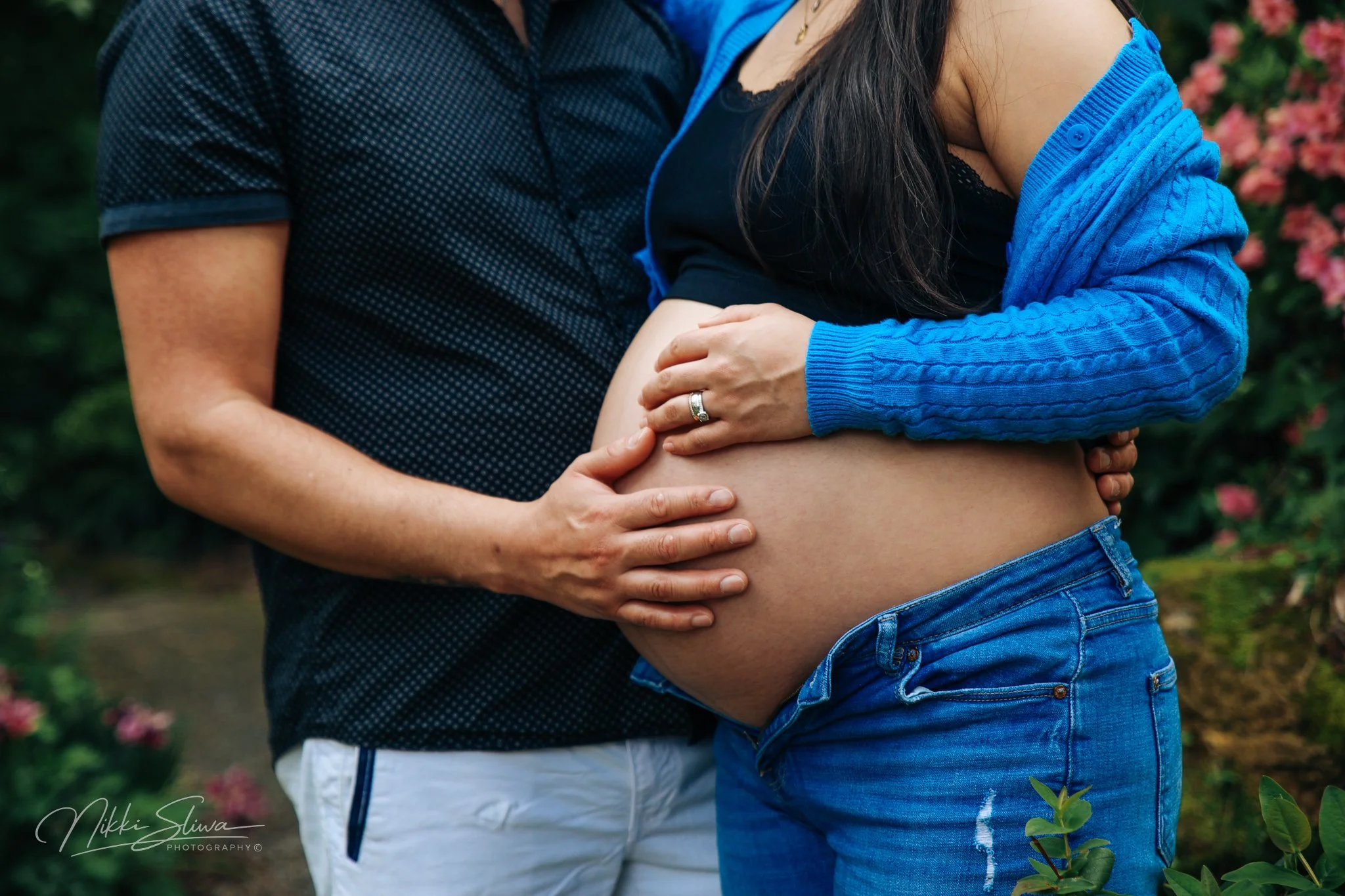 A pregnant woman with long dark hair in a black crop top and blue cardigan hugging her belly, with a ring on her finger, standing outdoors with pink flowers in the background, accompanied by someone in a dark patterned shirt and white pants, their ha