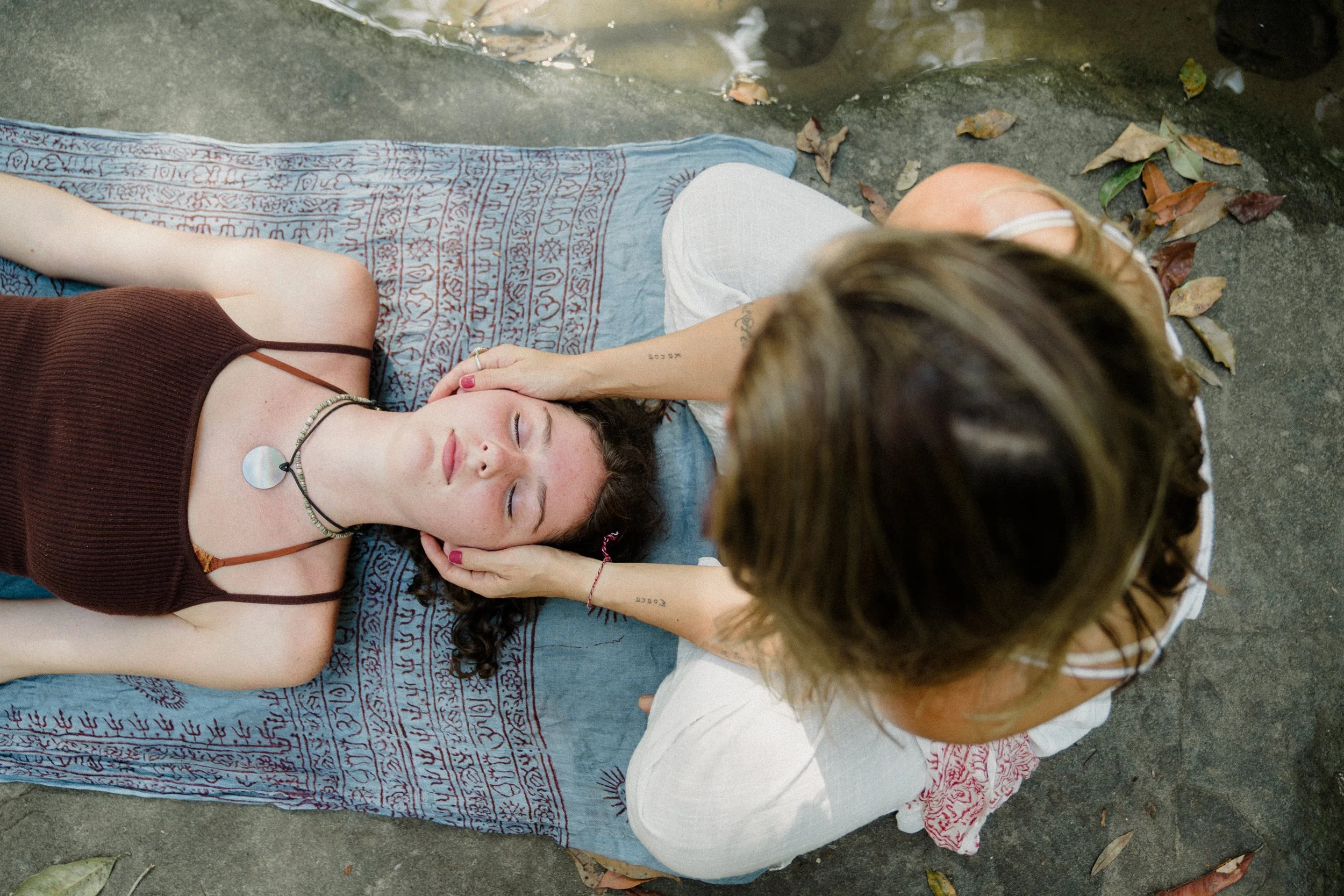 A person with dark hair, lying on a mat outdoors with eyes closed, receiving a head massage from another person with light brown hair. The person lying down has a necklace and wears a brown top, with a tattoo on their arm. The setting includes fallen leaves and a stone surface.