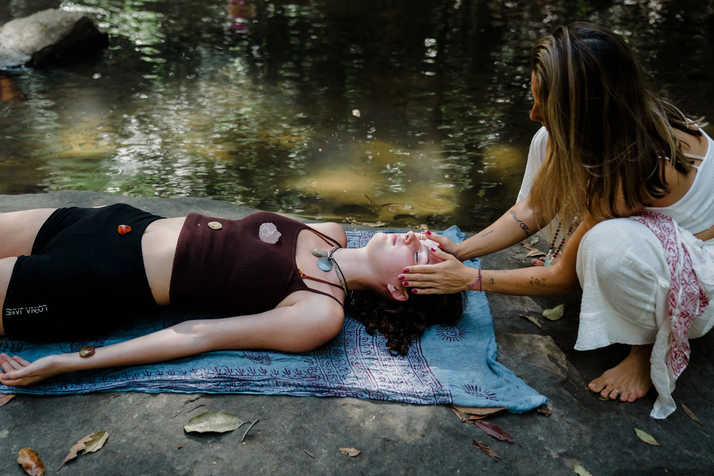 A woman lying on a towel near a river receiving a crystal healing treatment from another woman kneeling beside her.