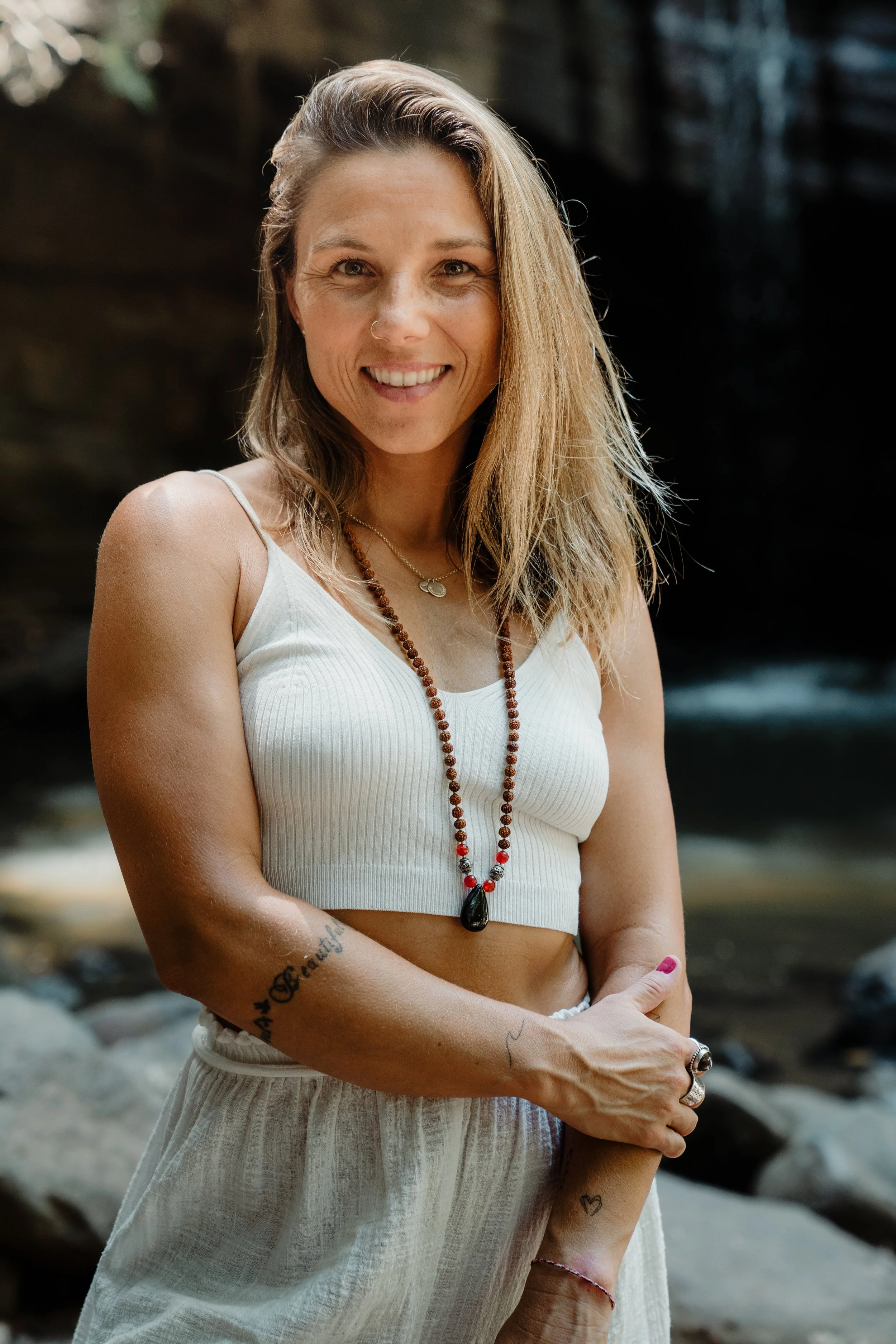 A woman with long, wavy blonde hair smiling outdoors, wearing a white crop top, white pants, and a beaded necklace, with tattoos on her arm, in front of a rocky background