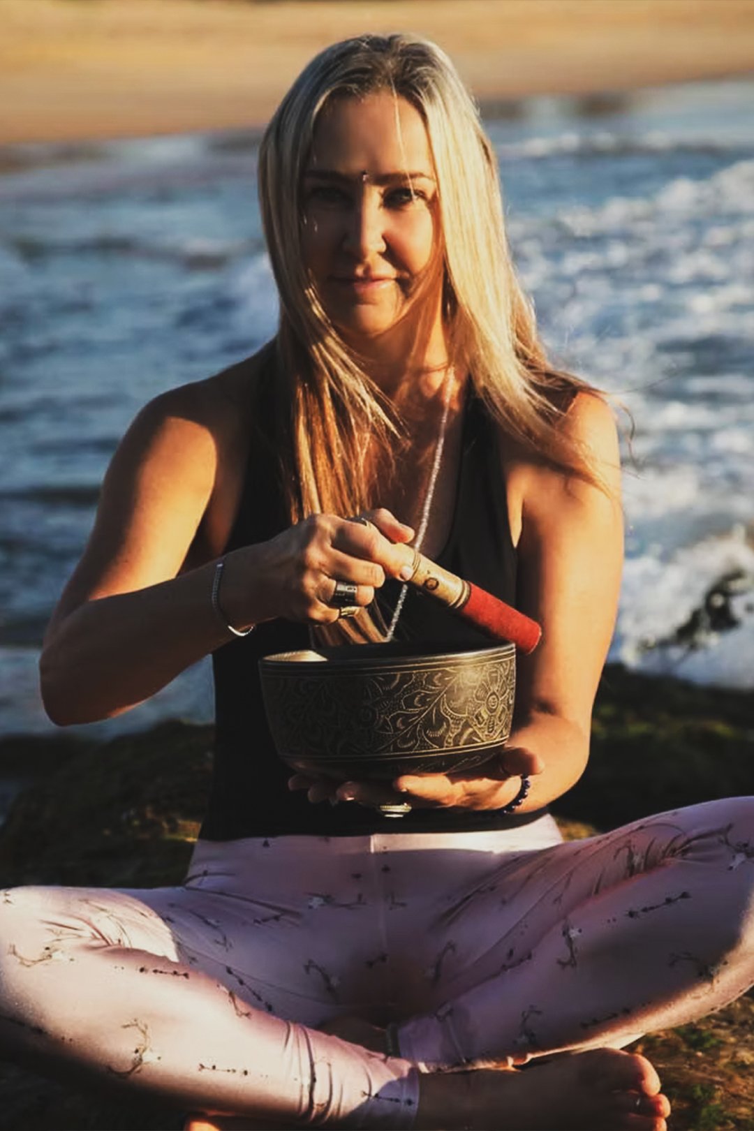 A woman sitting cross-legged on the beach during sunset, holding a singing bowl in one hand and a mallet in the other, with ocean waves in the background.