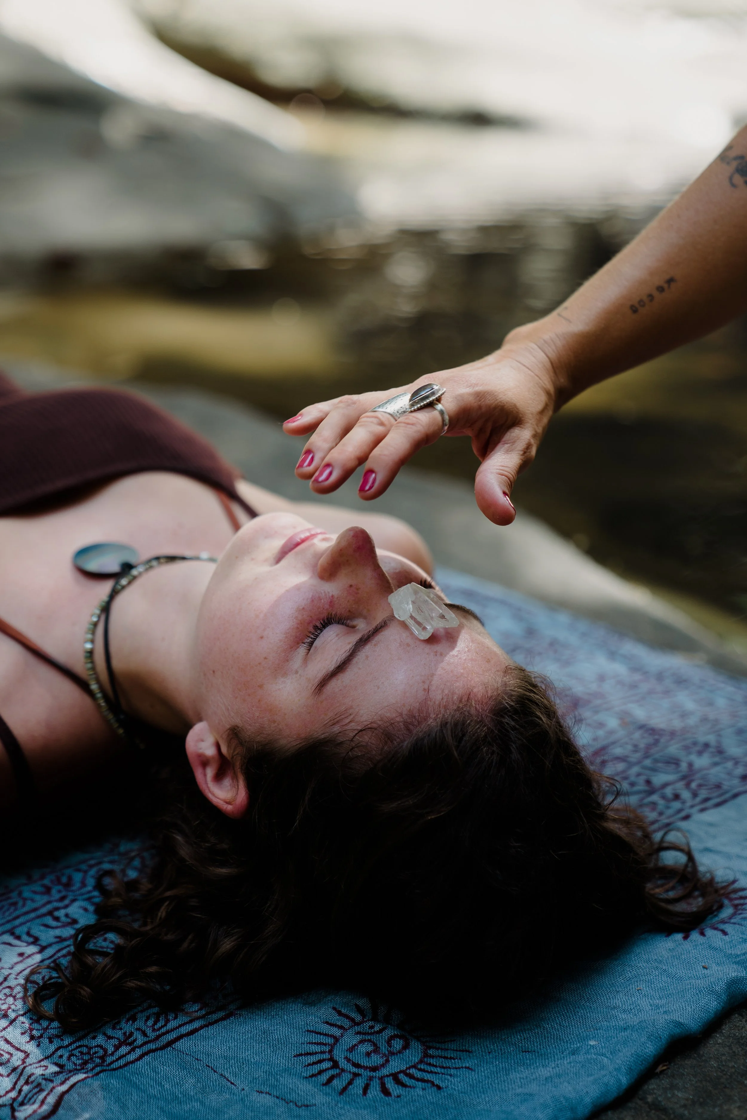 A woman lying on a patterned cloth outdoors, with a small crystal on her nose, receiving a gentle touch from another person.