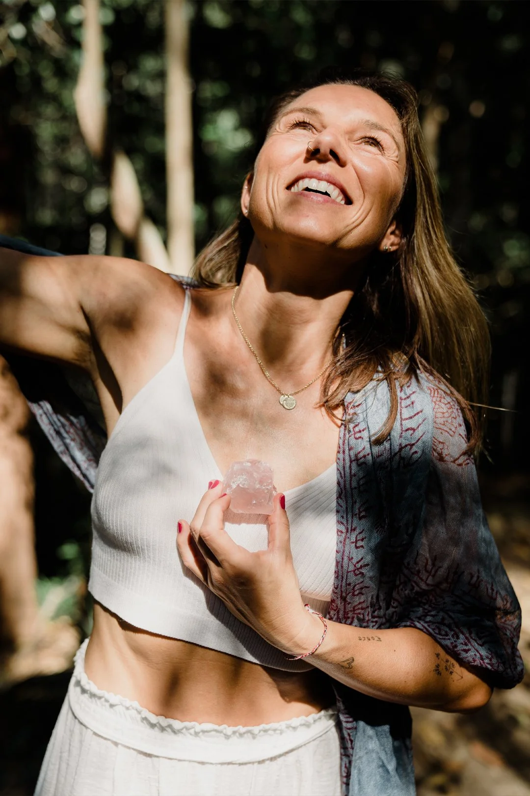 A woman in a white tank top holding a piece of crystal against her chest, smiling and looking up in a forest setting with sunlight filtering through trees.