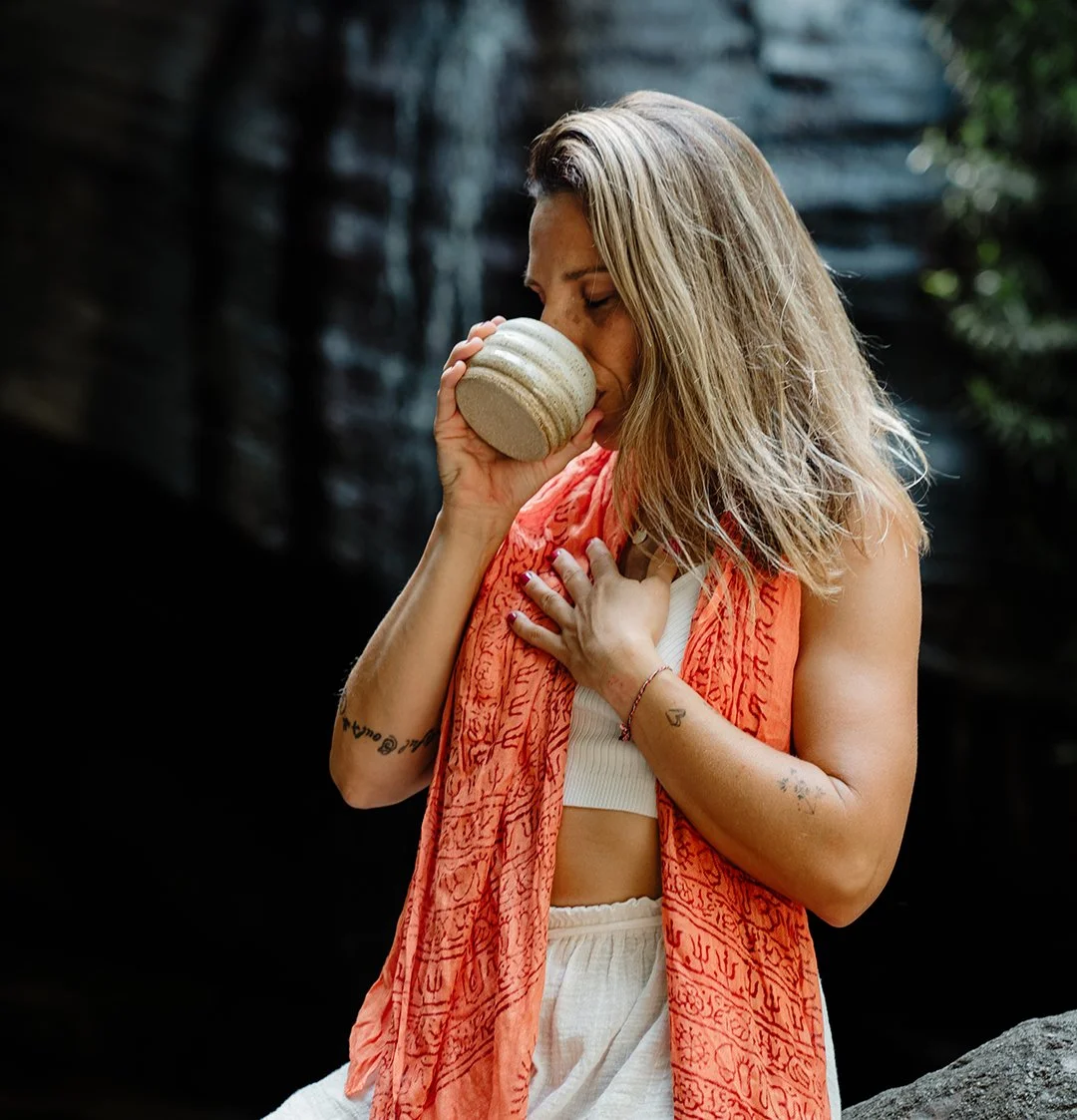 A woman with blonde hair, wearing a white top and an orange scarf, is holding a beige mug close to her face with her eyes closed, standing outdoors with a dark background and some greenery.