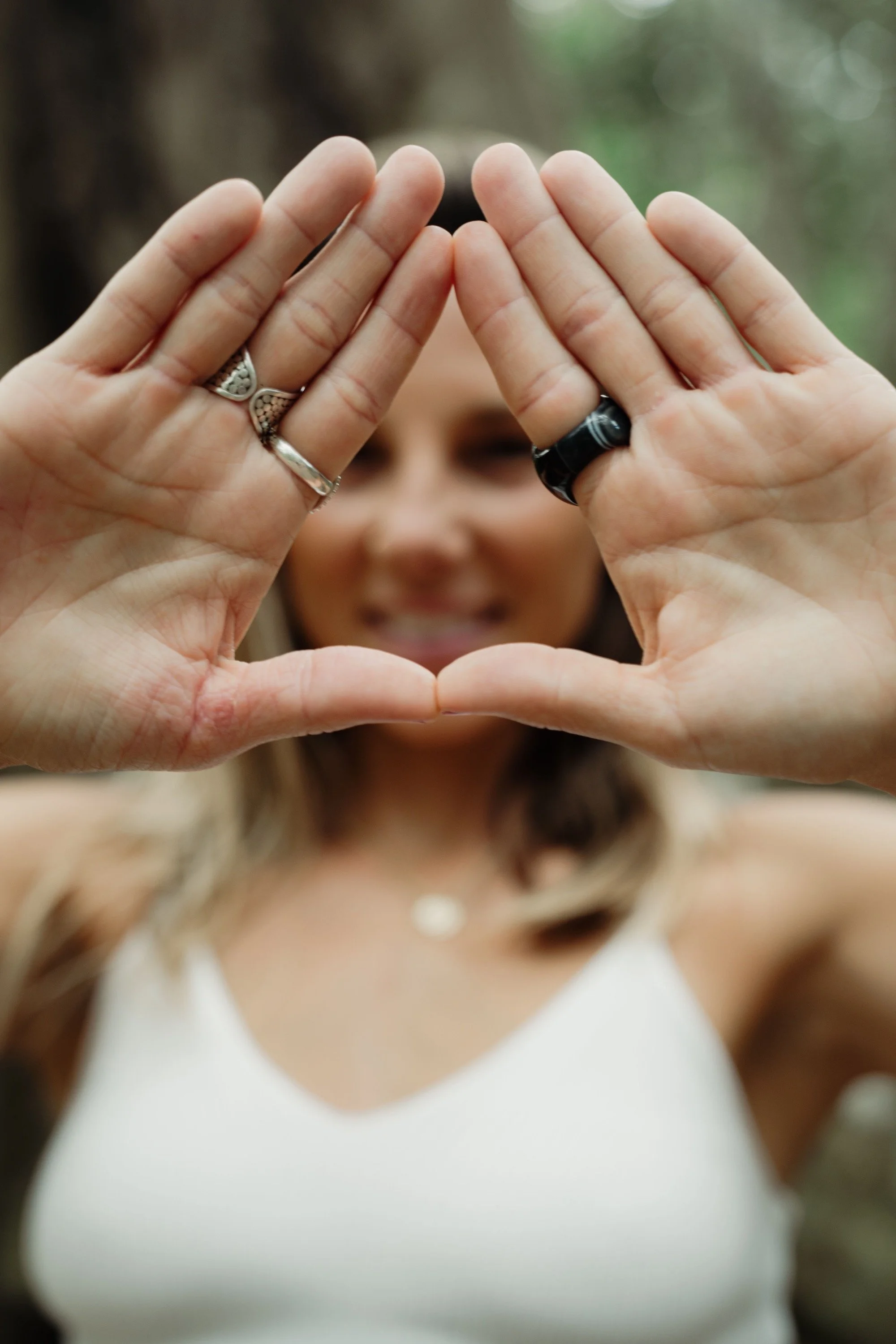 Person smiling outdoors, making a heart shape with hands in front of face, focusing on hands and rings.