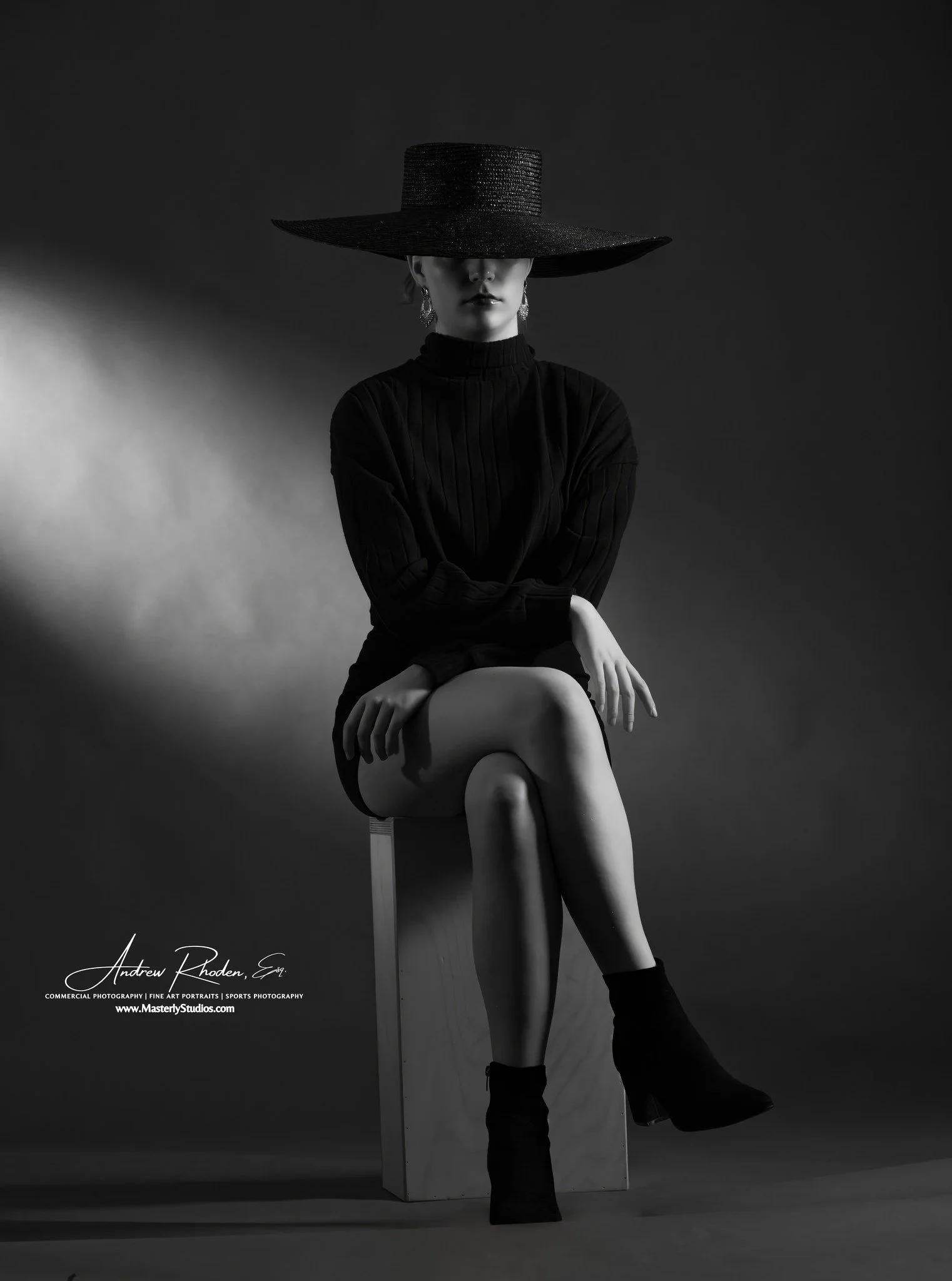 A black-and-white studio portrait of a woman sitting on a cube, wearing a wide-brimmed hat that obscures her eyes, a long-sleeved top, and ankle boots, with her legs crossed.