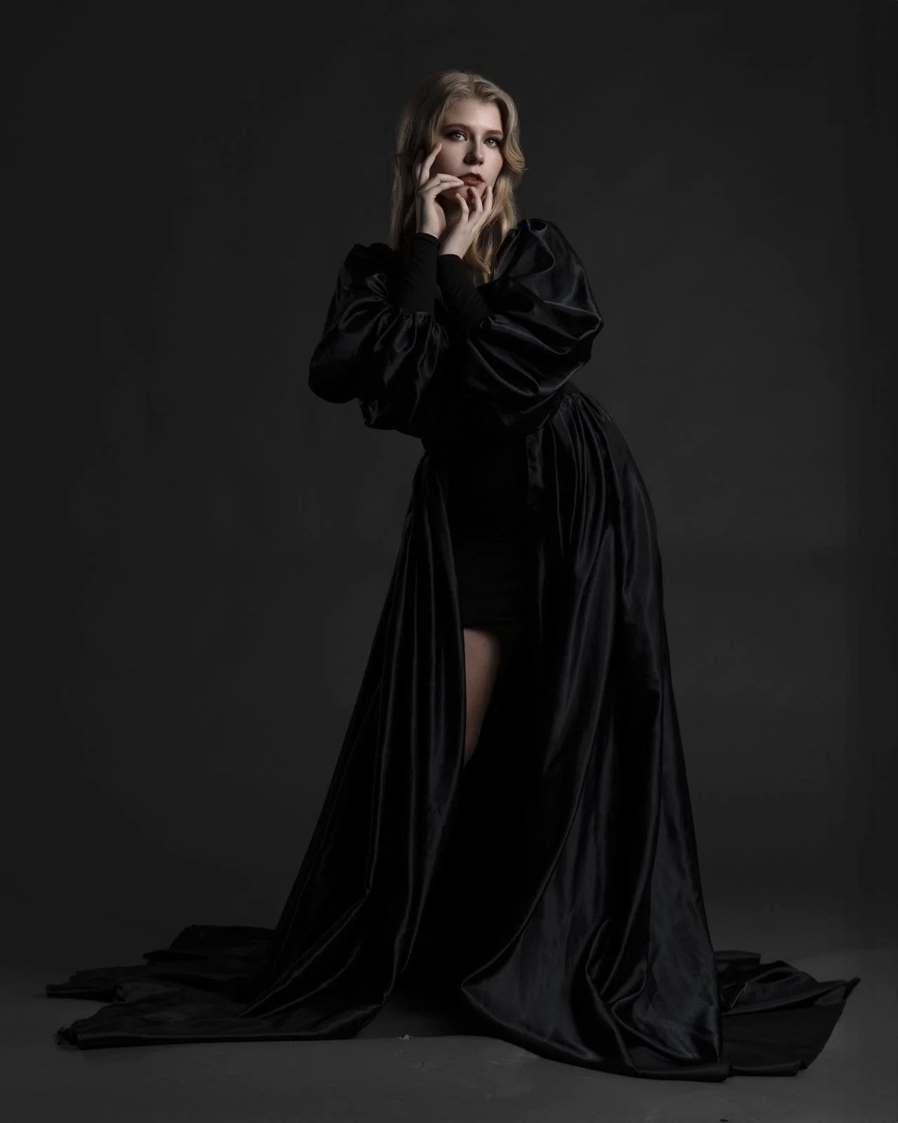 Addison Skyler modeling in a long, shiny black dress with puffed sleeves, standing against a dark background, with one leg slightly raised and her hands near her face.