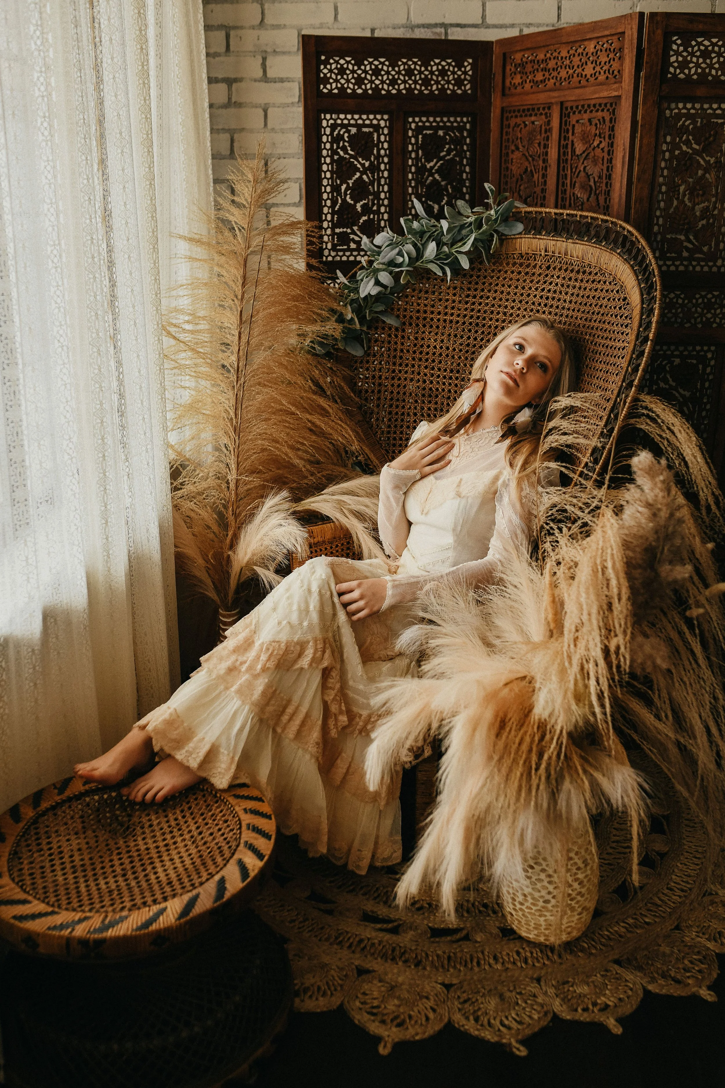 A young girl with long blonde hair, dressed in a vintage cream-colored dress, sitting on a wicker chair surrounded by tall dried pampas grass and greenery, in a warmly lit room with lace curtains and wooden folding screen.