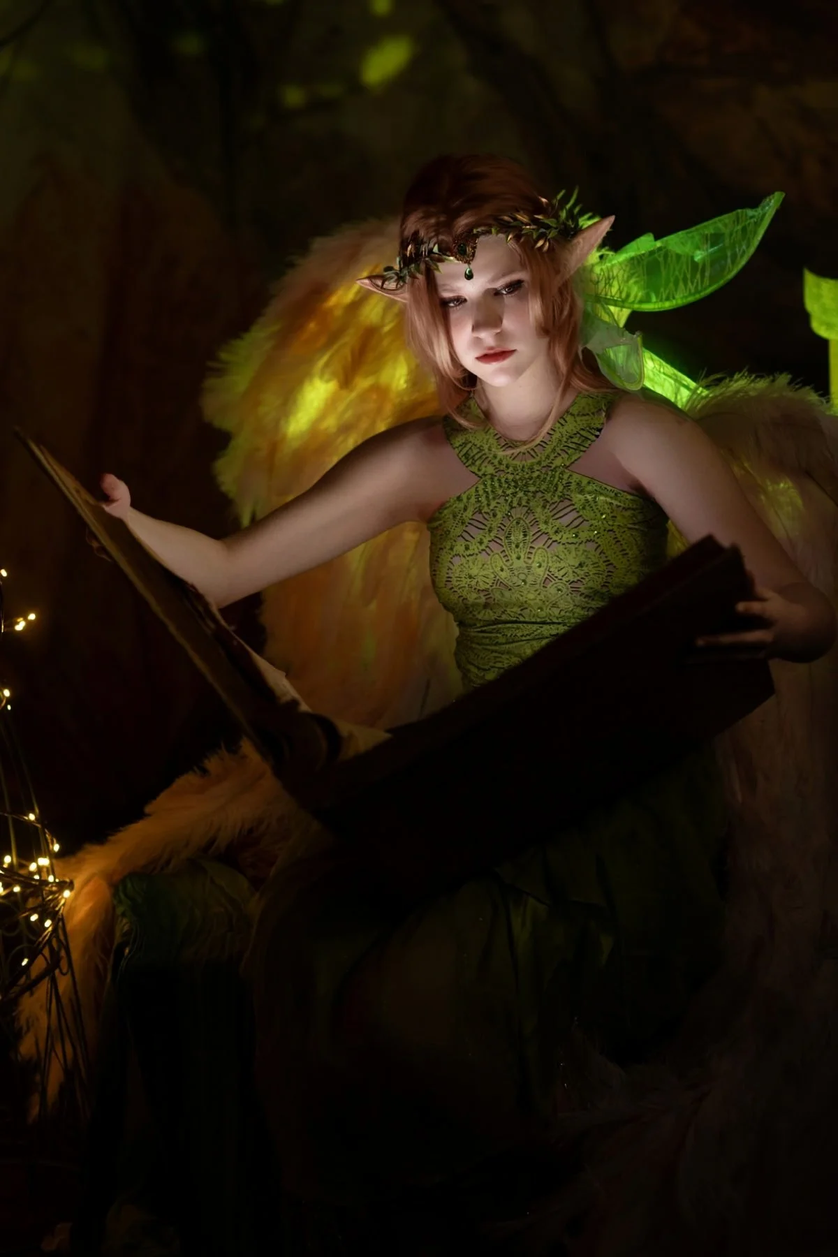 Addison Skyler in fantasy photo shoot. A fantasy elf woman with leaf-shaped glowing green wings, pointed ears, and a leafy headpiece, reading a book in a dimly lit environment with fairy lights.