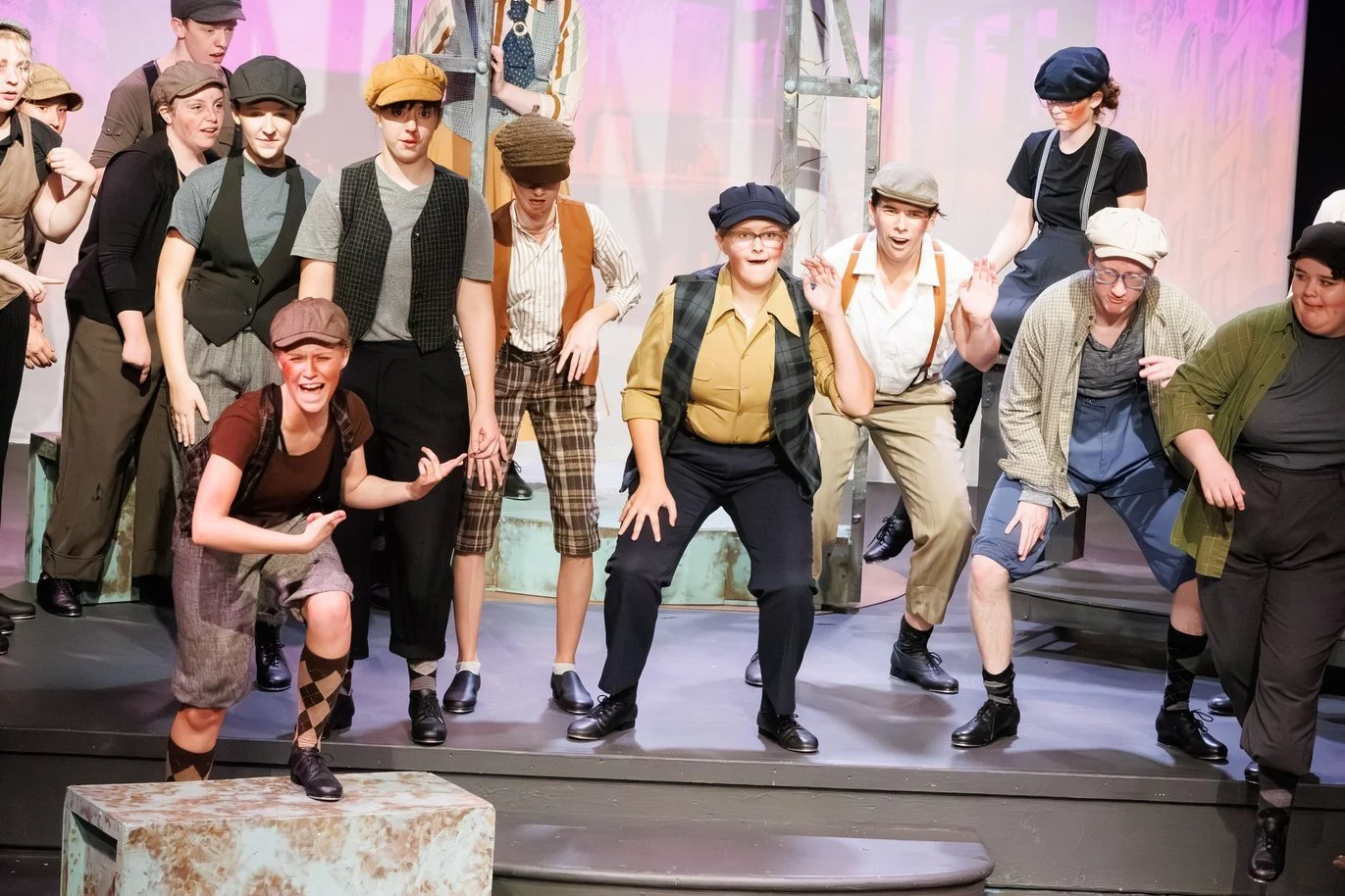 Addison Skyler dancing and performing on stage in Newsies a theatrical play, dressed in vintage clothing, some with caps and suspenders, with a scaffold in the background.