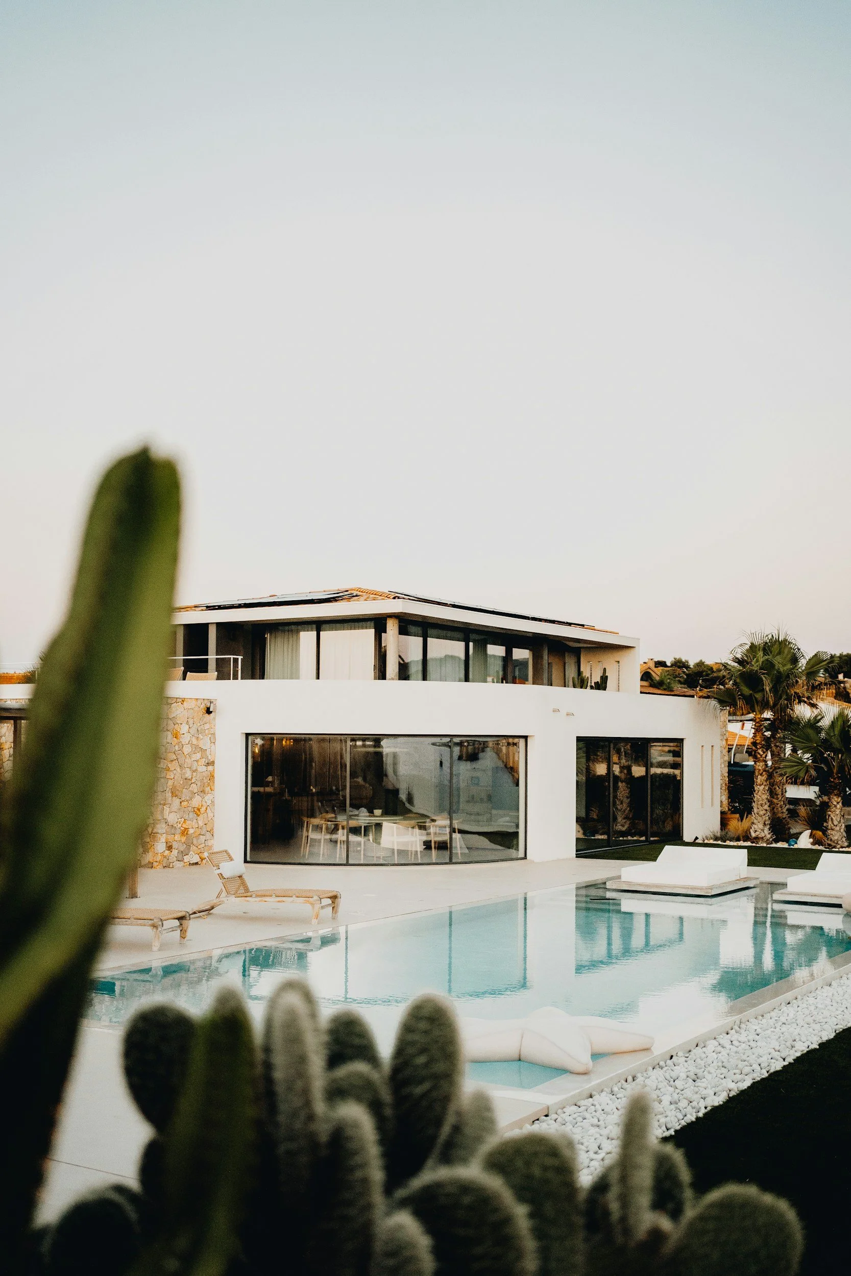 View of a modern house with large glass windows, a swimming pool with lounge chairs, and surrounding desert plants and cacti, during daytime.
