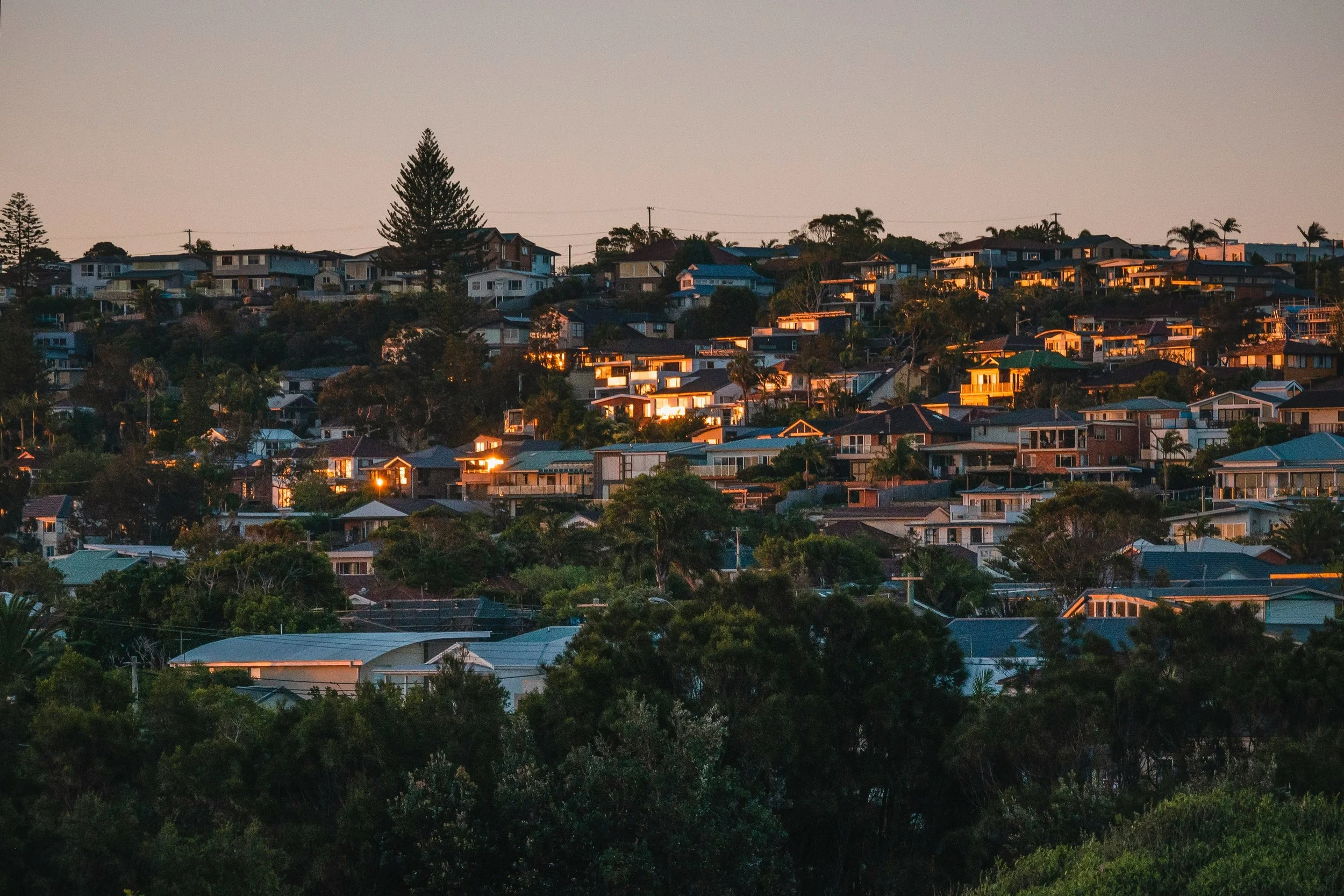 Sunset over a hillside neighborhood with houses and trees, glowing yellow and orange reflections on buildings.