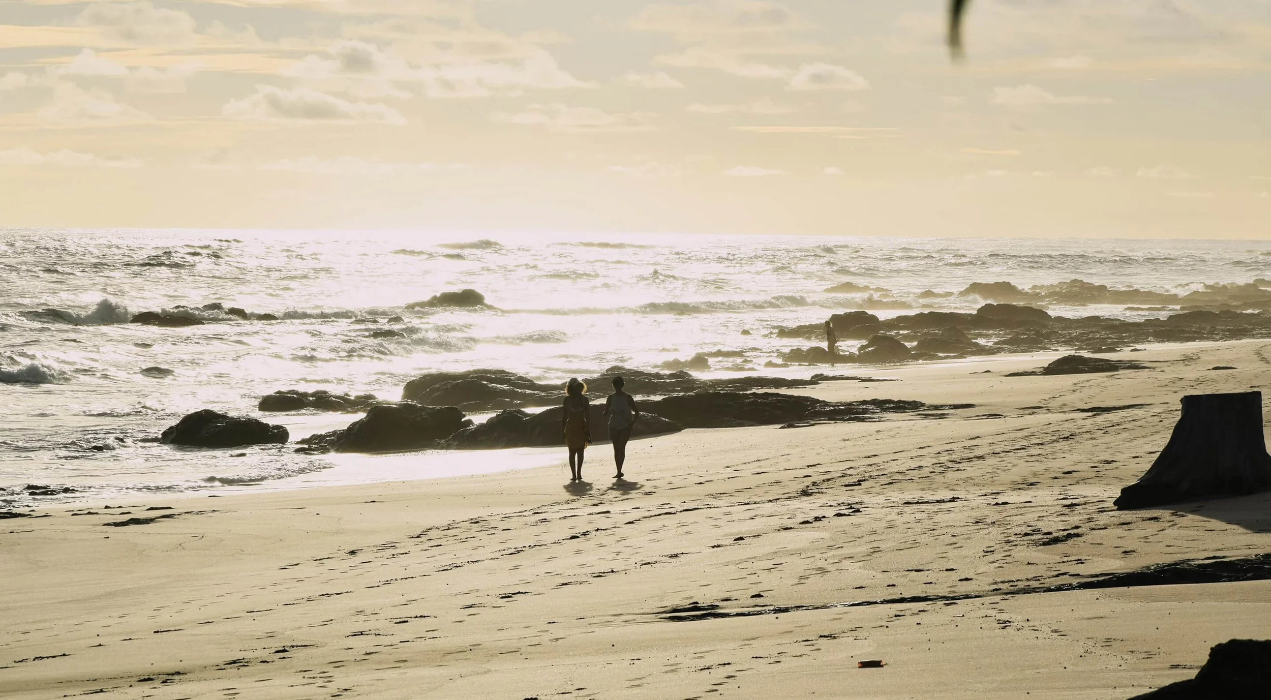 People walking along the beach near rocks with the ocean and a cloudy sky in the background during sunset or sunrise.