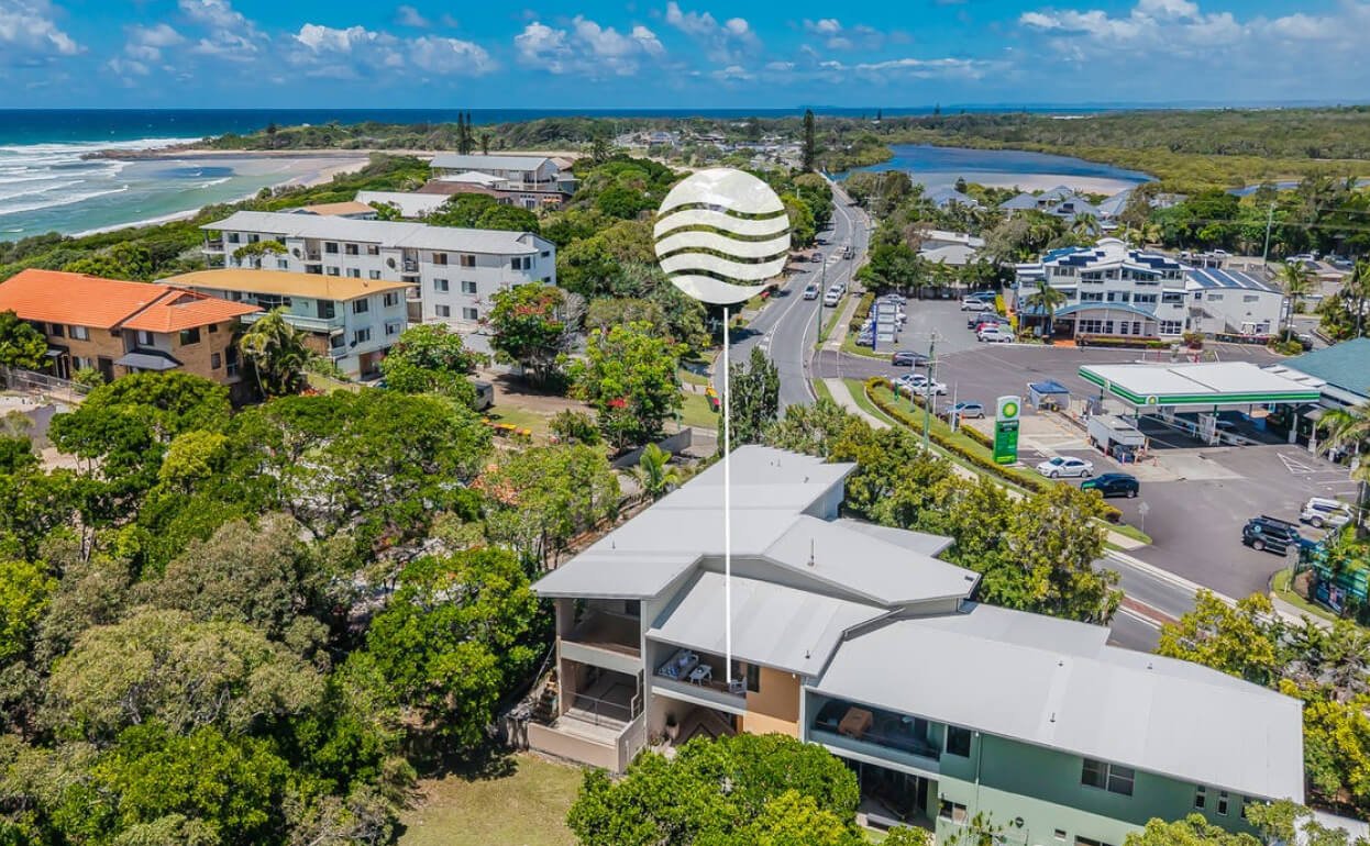Aerial view of a coastal residential area with a building featuring a large striped sphere on a pole, trees, a roadside with parked cars, and a distant ocean and sky.