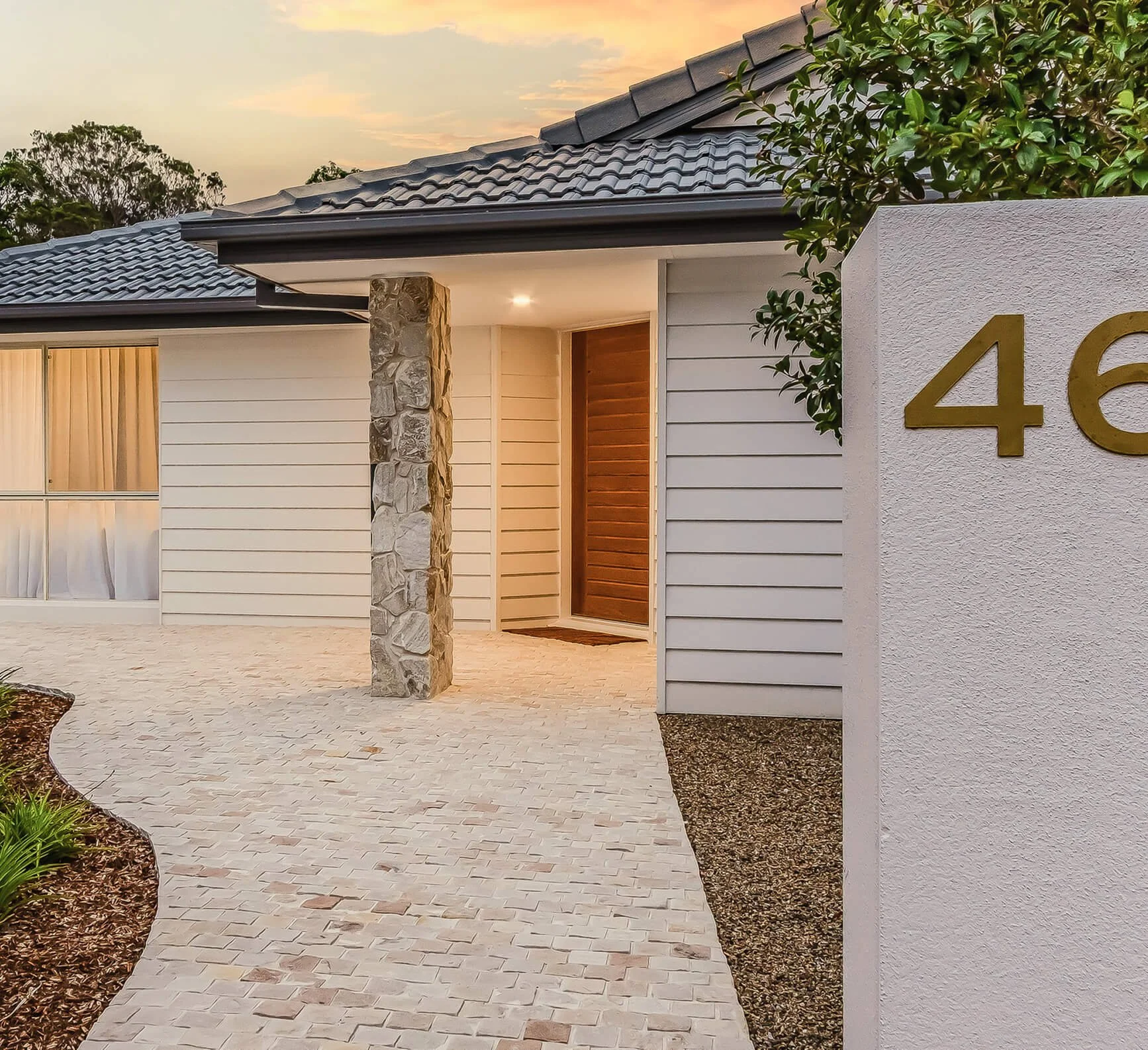 Modern house with a stone pillar at the entrance, white horizontal siding, a large window with curtains, and a wall with house number 46. Brick pathway and landscaped garden in front, trees visible in the background at sunset.