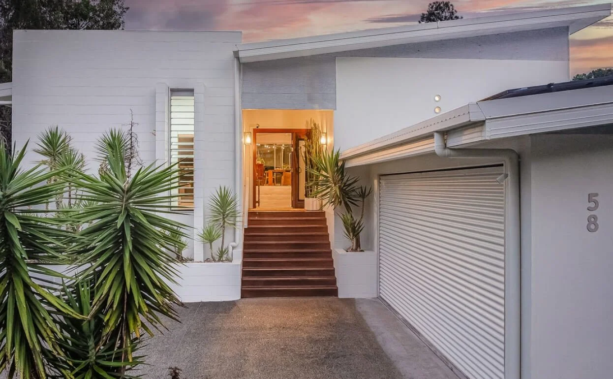 Modern white house with wooden stairs leading to a lit entryway, surrounded by green plants, at sunset.