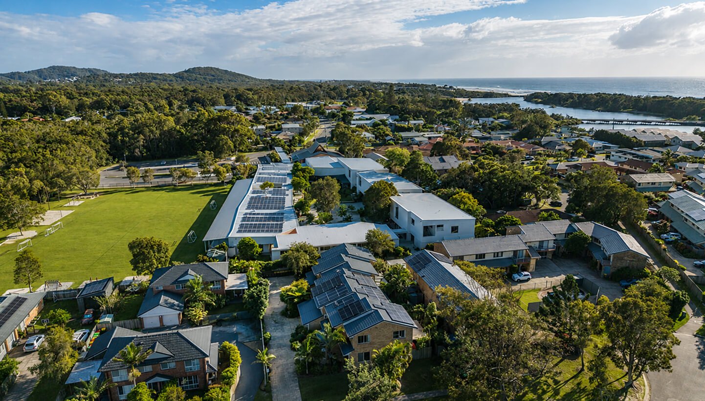Aerial view of a residential neighborhood with houses, a school with a large field and solar panels, surrounded by greenery and hills, near a body of water under a partly cloudy sky.