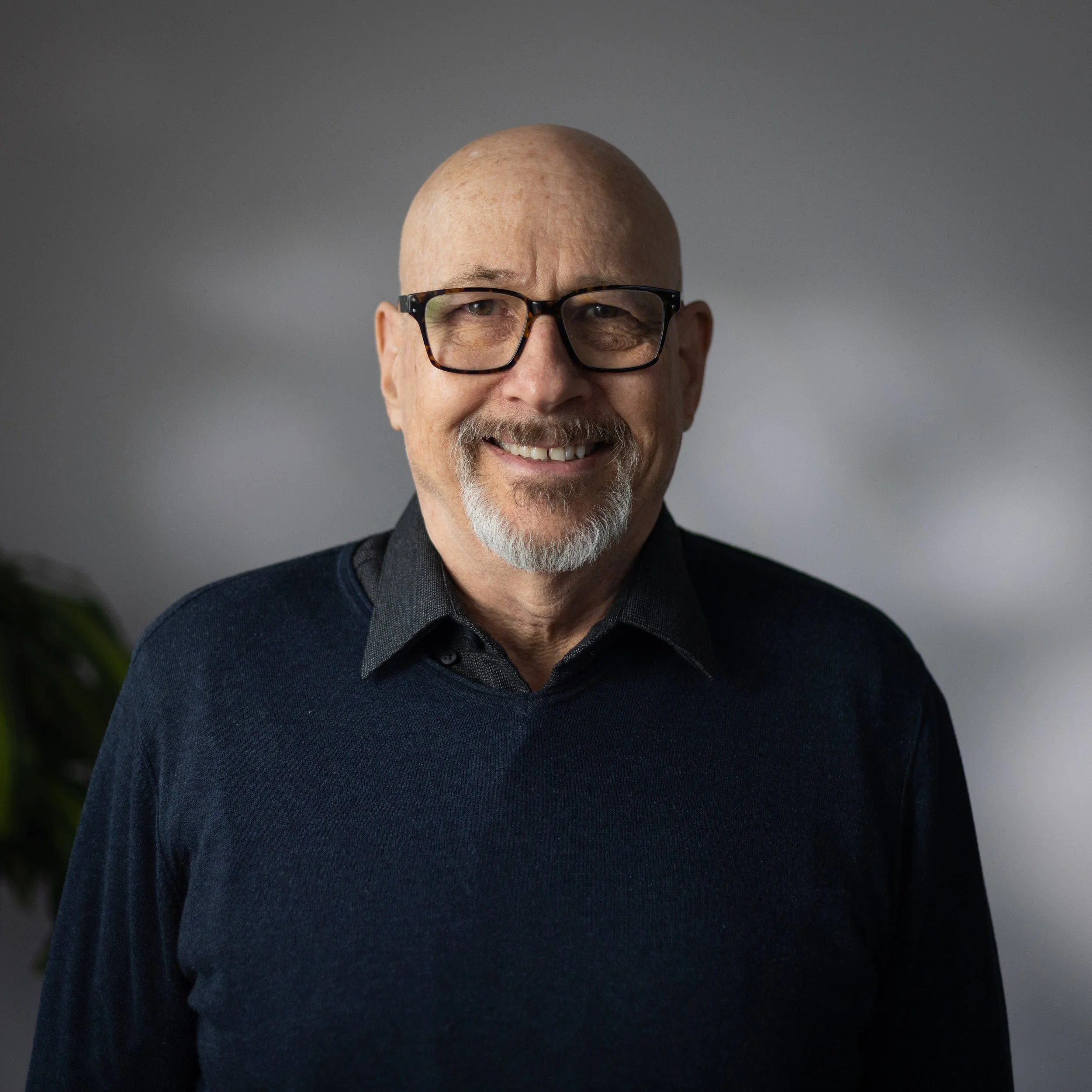 Portrait of a smiling older man with glasses, light beard, and bald head, wearing a dark sweater over a collared shirt, in front of a plain gray background.