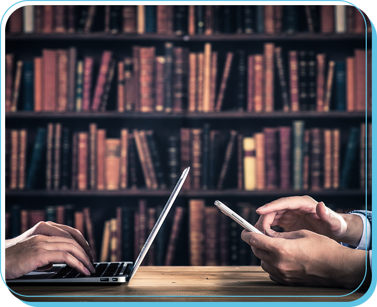 Two people using a laptop and a smartphone at a wooden table with a bookshelf filled with books in the background.