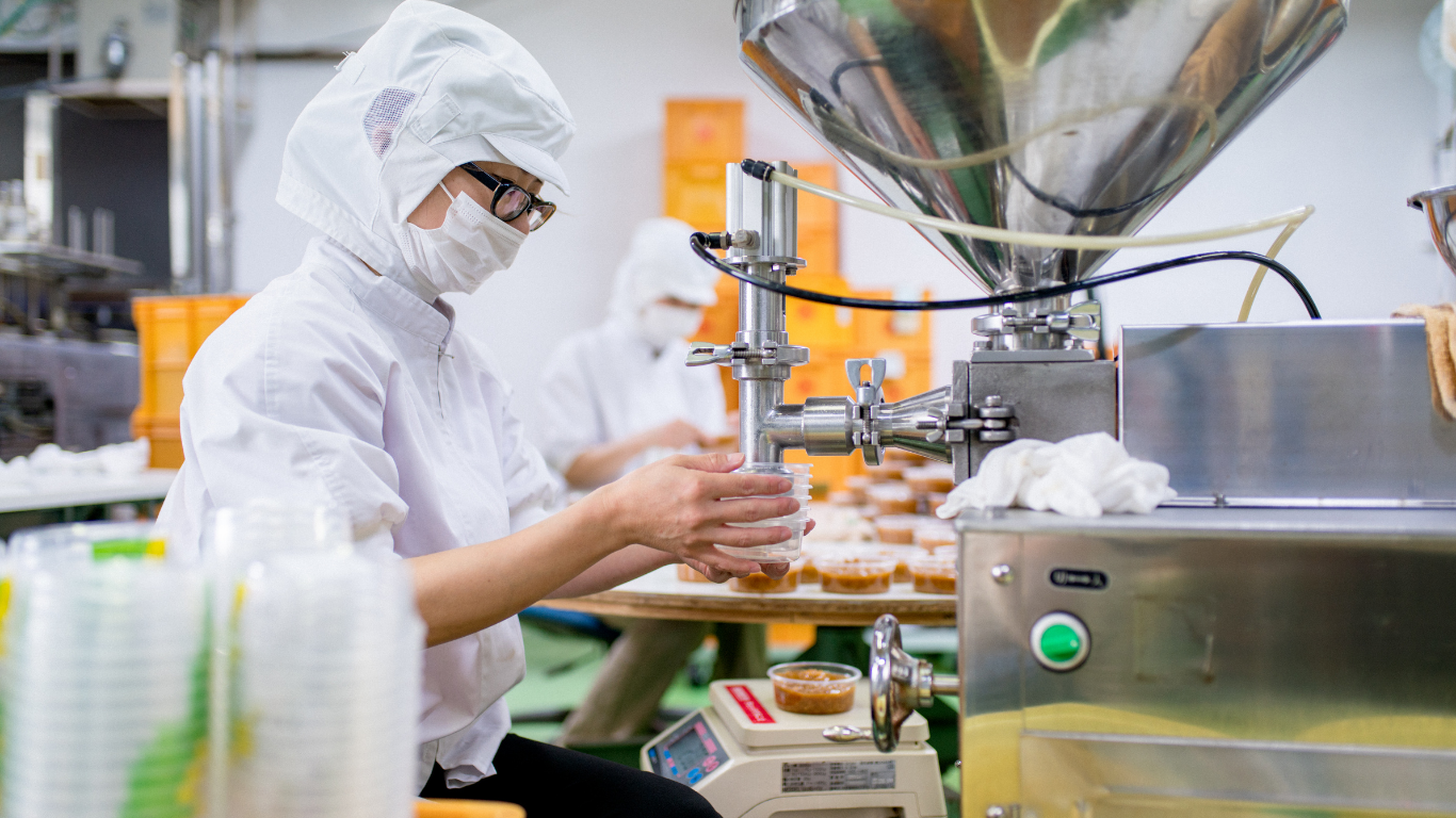 A worker wearing a white protective suit, mask, and glasses uses machinery to fill small containers with a product in a food production facility.
