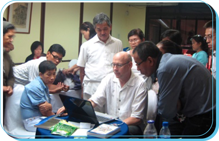 A group of people gathered around a man working on a laptop at a meeting or workshop in a conference room.