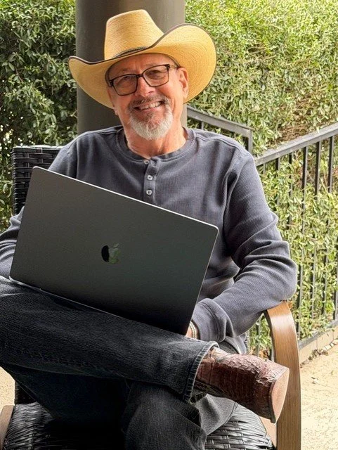 A man with glasses and a beard wearing a straw hat and a gray shirt, sitting outside with a closed MacBook in his lap, smiling.