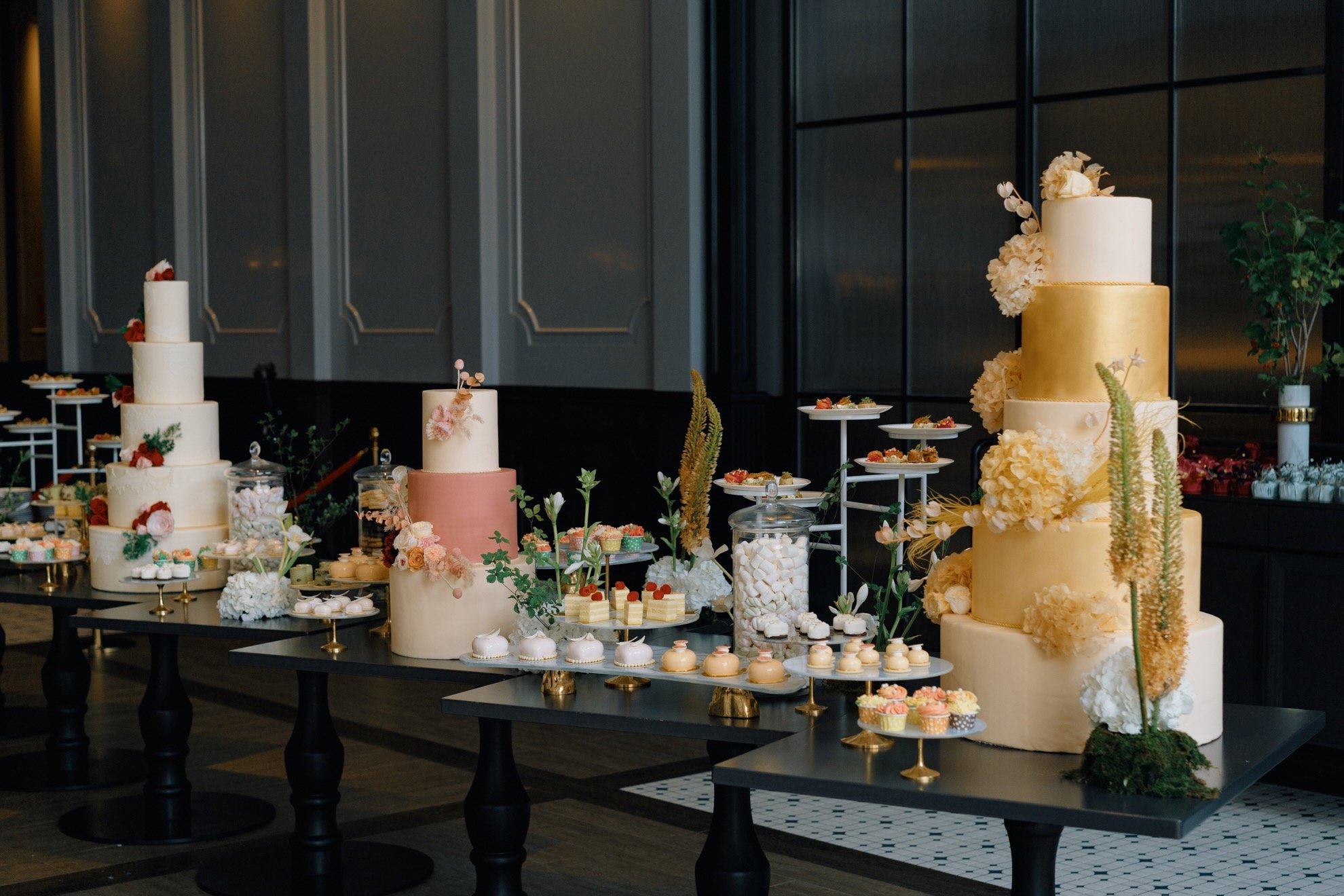 Elegant dessert display featuring multiple tiered cakes and assorted sweets on a black table, decorated with flowers and greenery, in a sophisticated event setting.