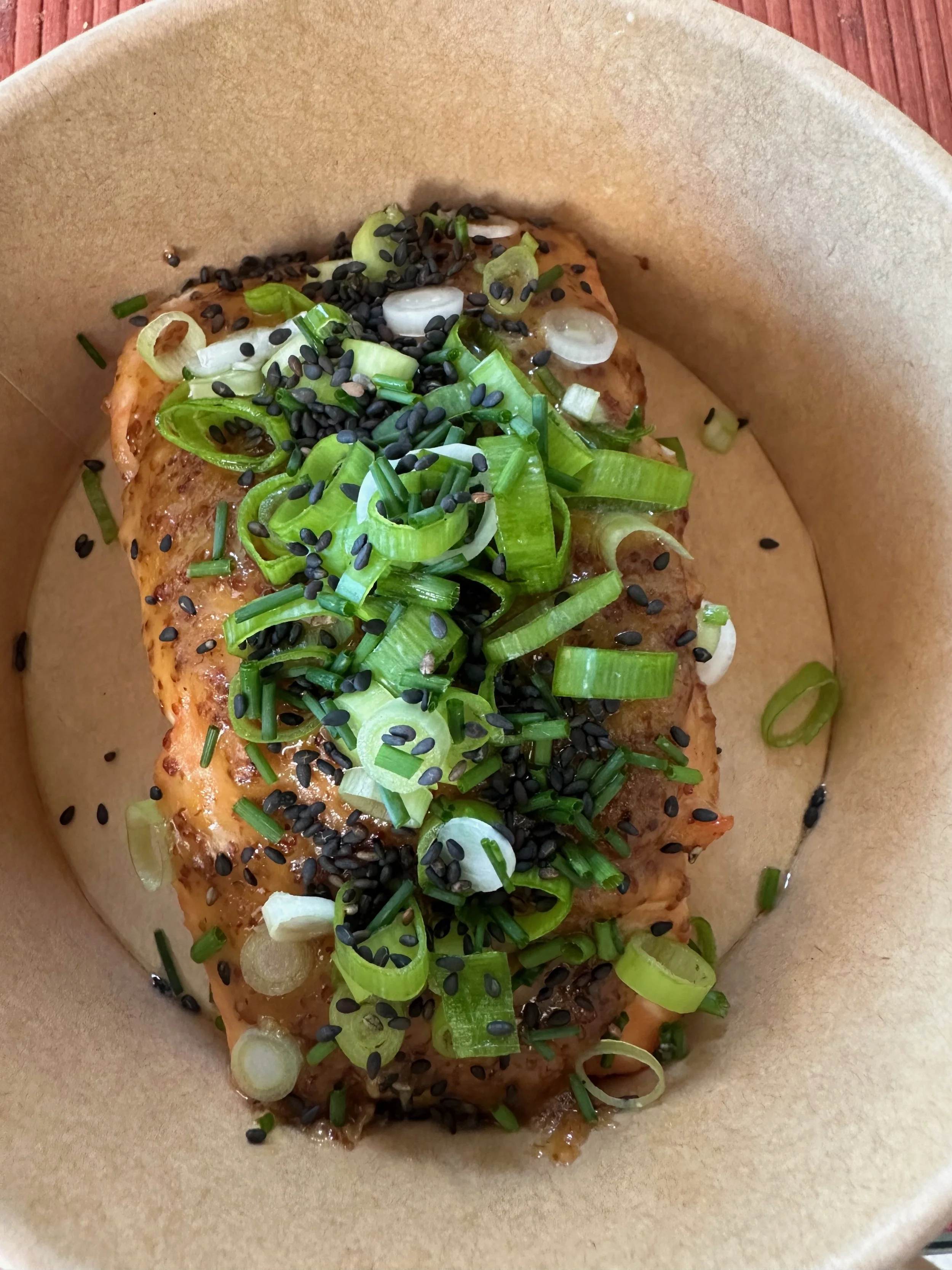 A close-up of a cooked dish topped with sliced green onions and black sesame seeds, served in a brown paper food tray.