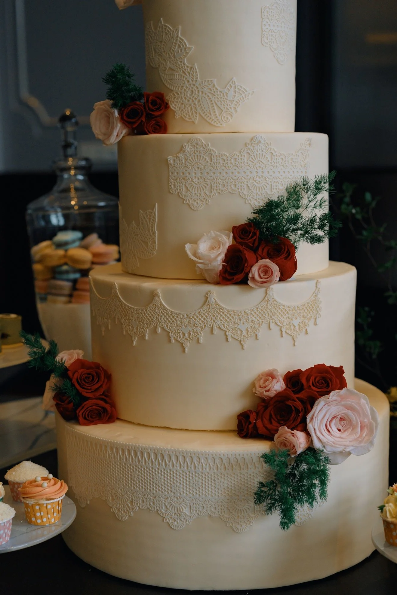 A four-tier white wedding cake decorated with red and pink roses, green foliage, and intricate lace-like patterns, with cupcakes nearby.