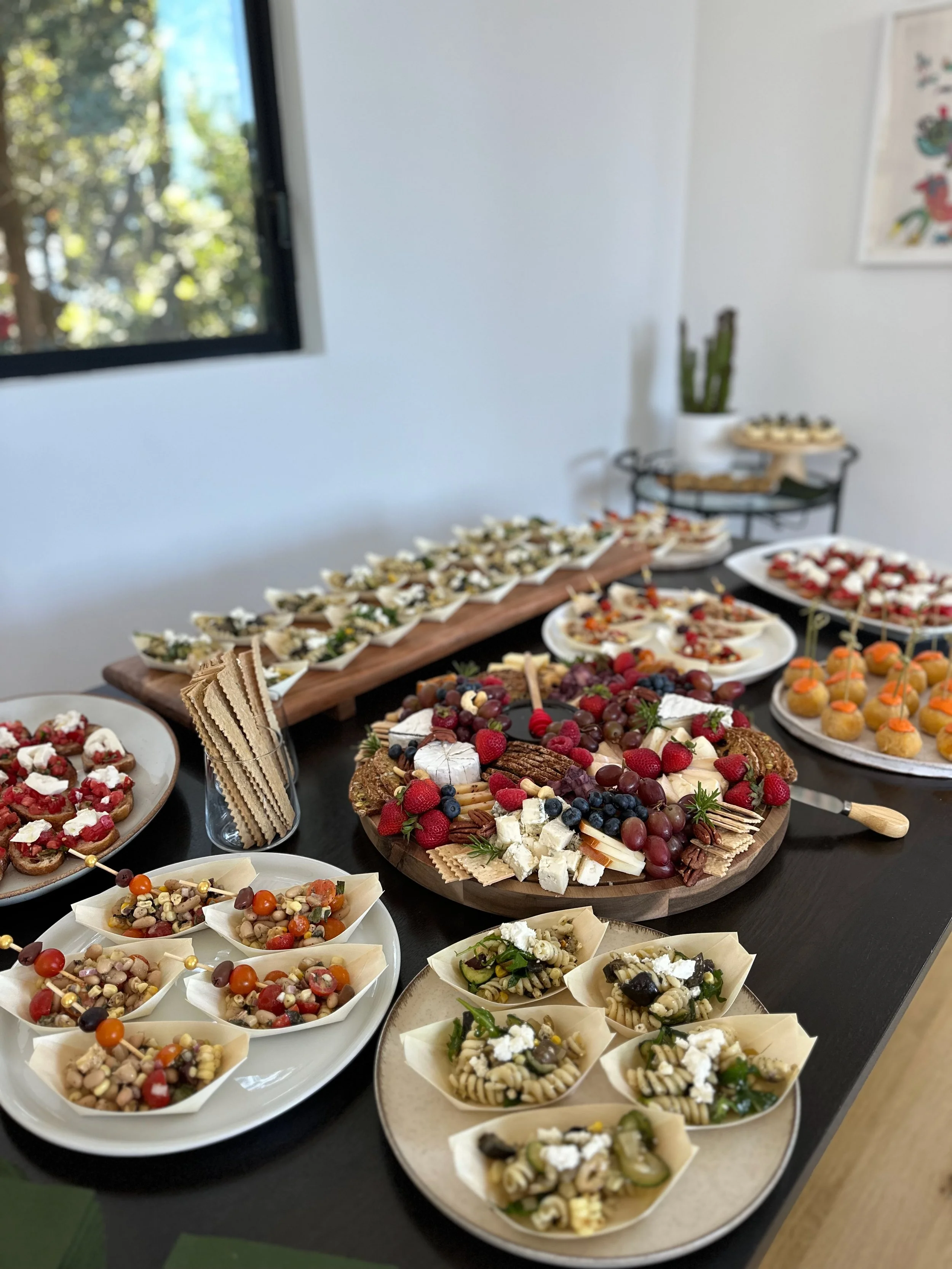 A table filled with various colorful appetizers, cheeses, and fruits, including grape, cucumber, and berry garnished dishes, set in a bright room with a window and a cactus plant in the background.