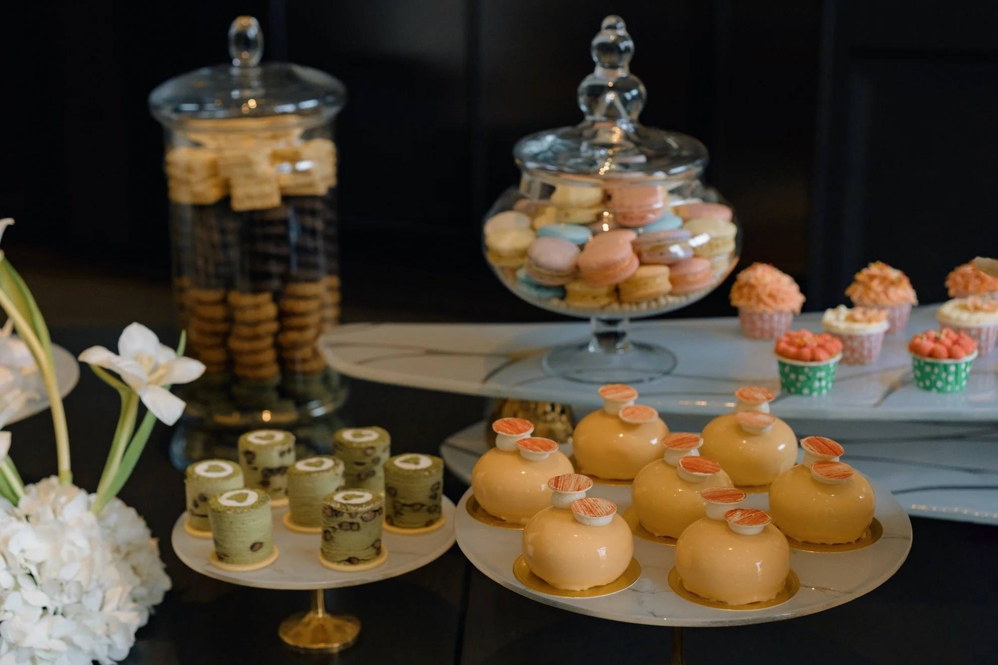 Decorative display of assorted desserts and sweets, including macarons, cupcakes, and petite cakes on tiered trays and glass jars at a celebration or event.