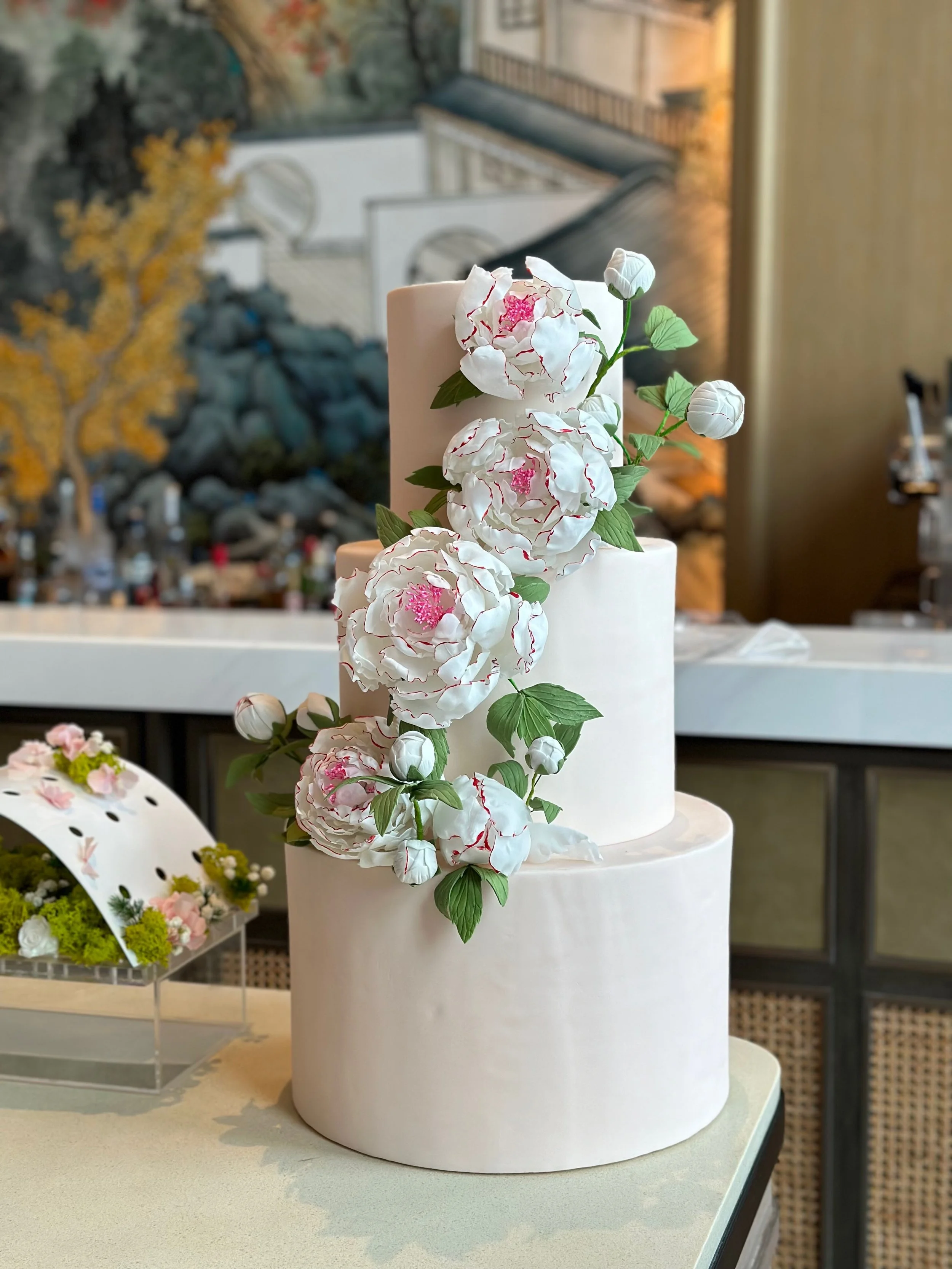 A two-tiered white wedding cake decorated with cascading white flowers and green leaves, placed on a table with a blurred background of an indoor setting.