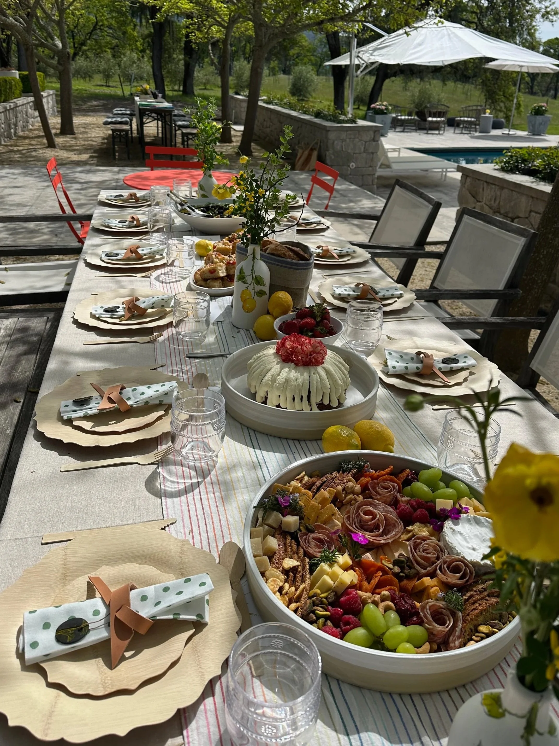 Outdoor dining table set for a celebration with various dishes including a cheese platter, desserts, and flowers, shaded by trees with a pool and patio in the background.