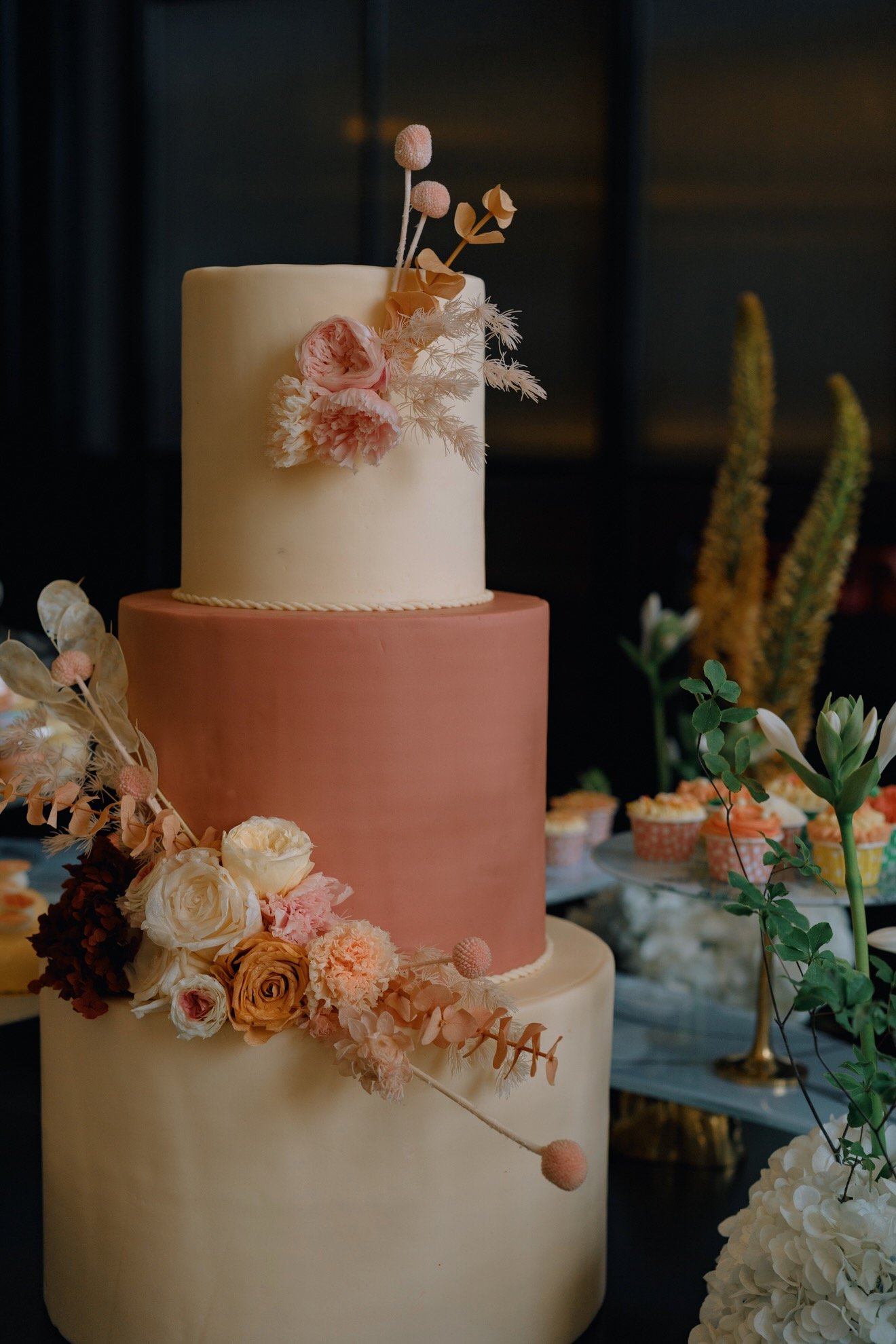 A three-tiered wedding cake decorated with pastel-colored flowers and dried foliage, with additional floral arrangements on a table in the background.