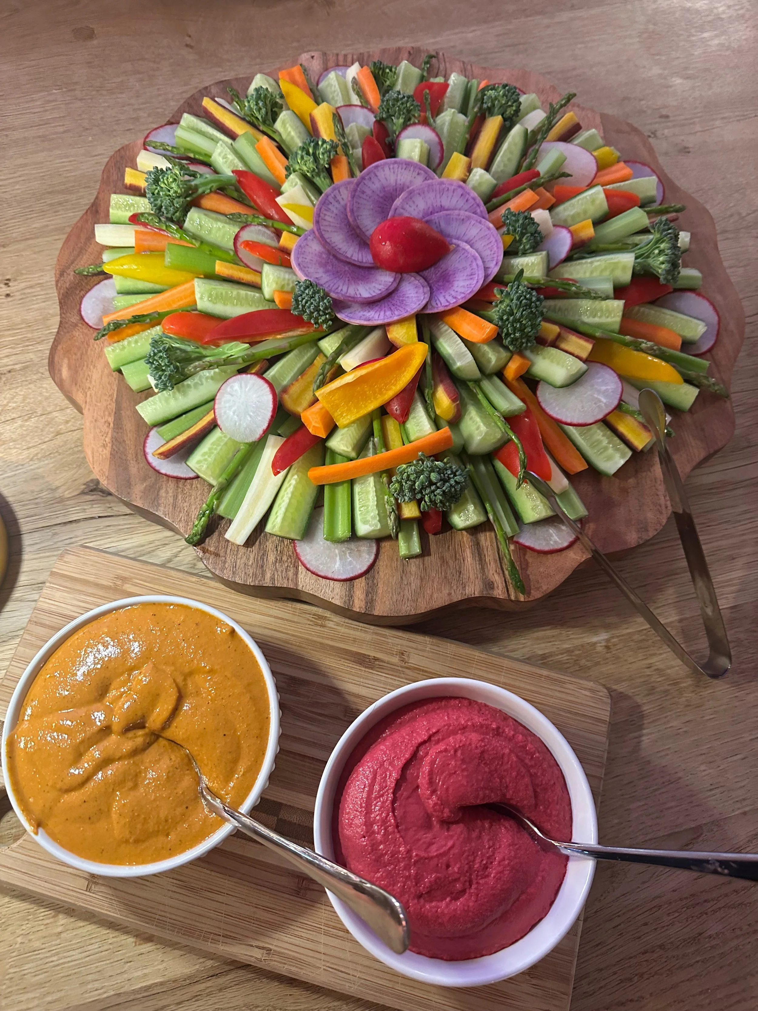 Colorful vegetable platter on a wooden serving board with two bowls of dip, one yellow and one pink, on a wooden table.