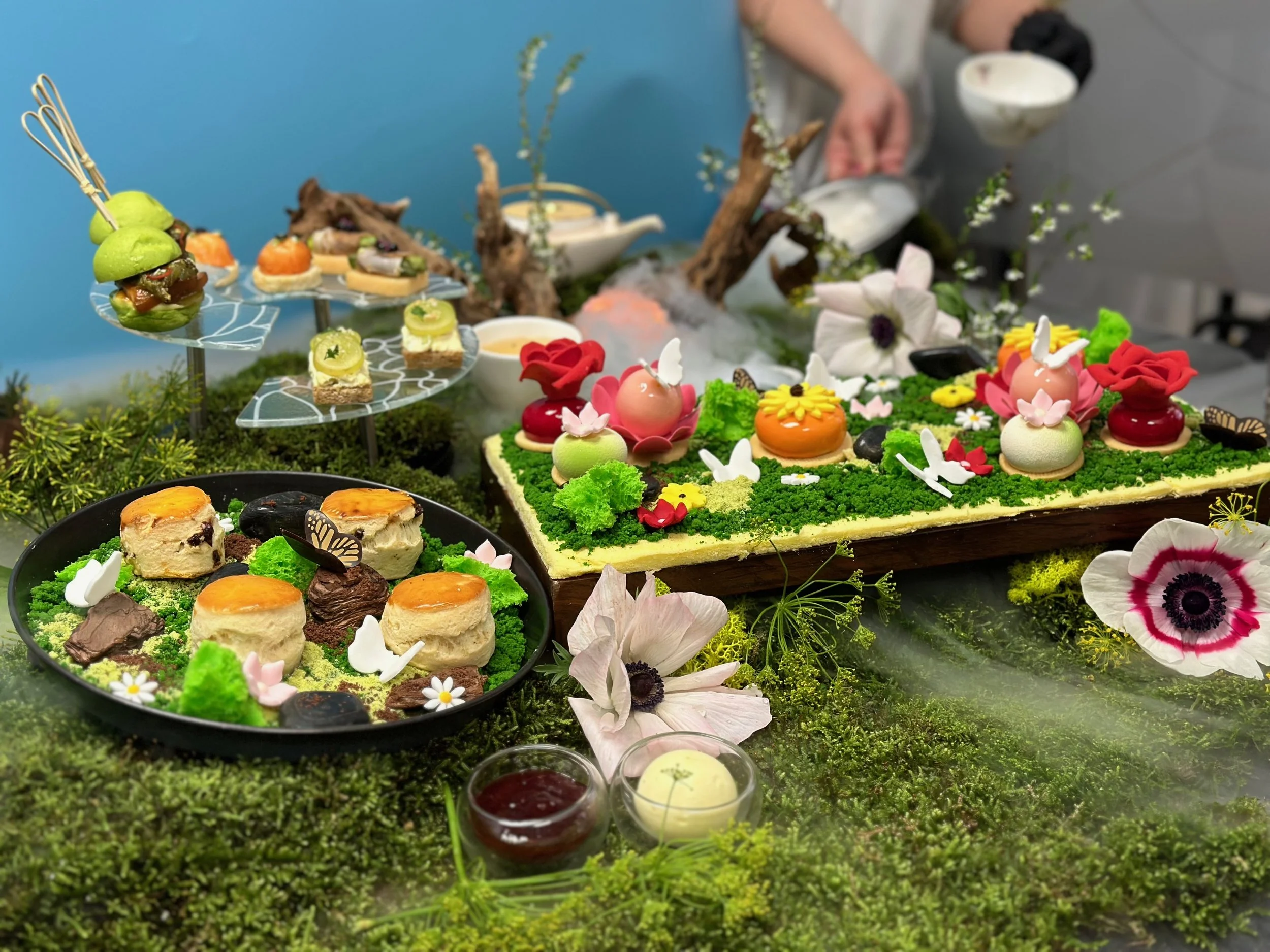 Assorted decorative desserts and pastries arranged on a moss-covered display with flowers, greenery, and a blue backdrop.