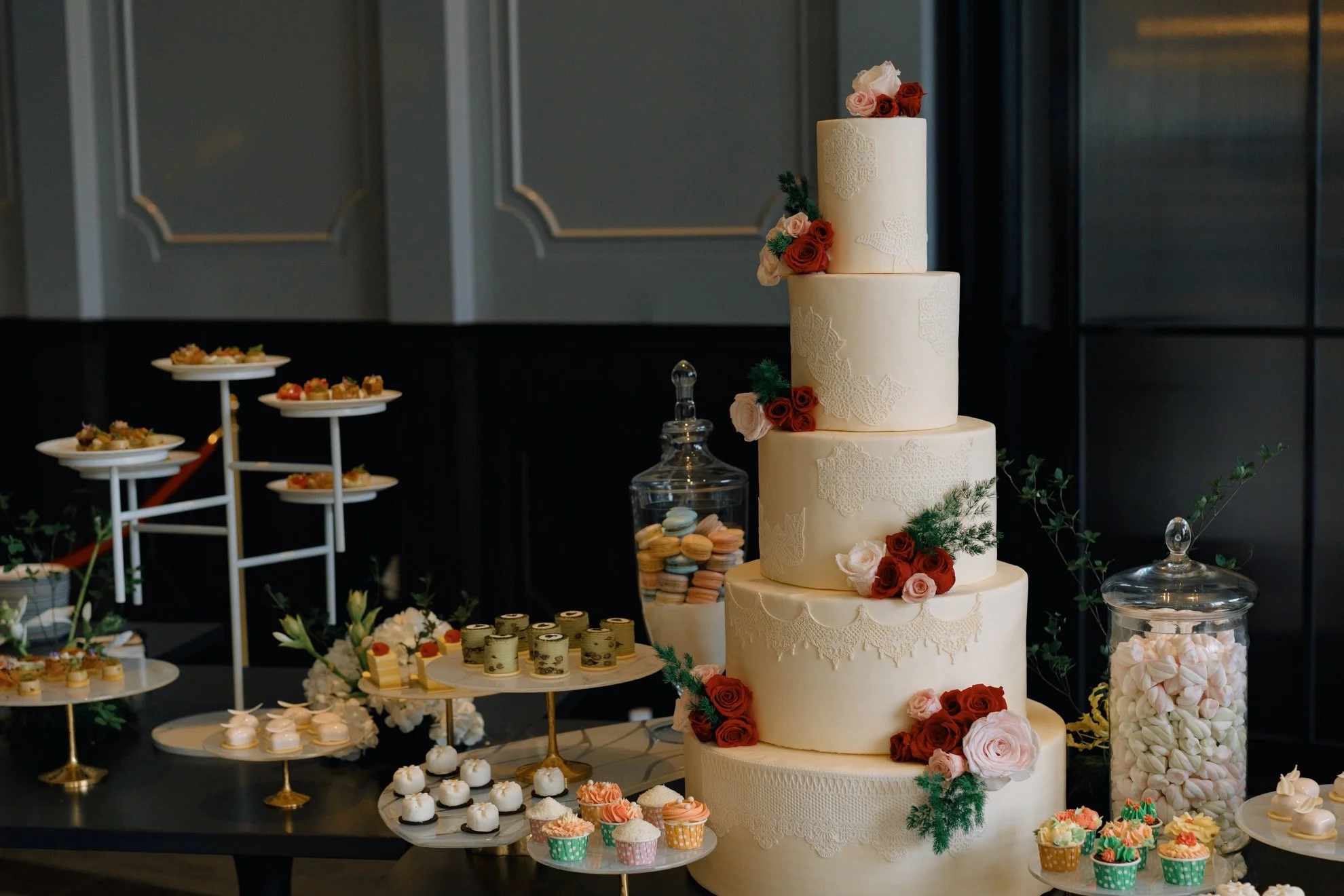 Four-tiered white wedding cake decorated with pink and red roses and greenery, surrounded by various desserts like cupcakes, macarons, and small pastries on a table.
