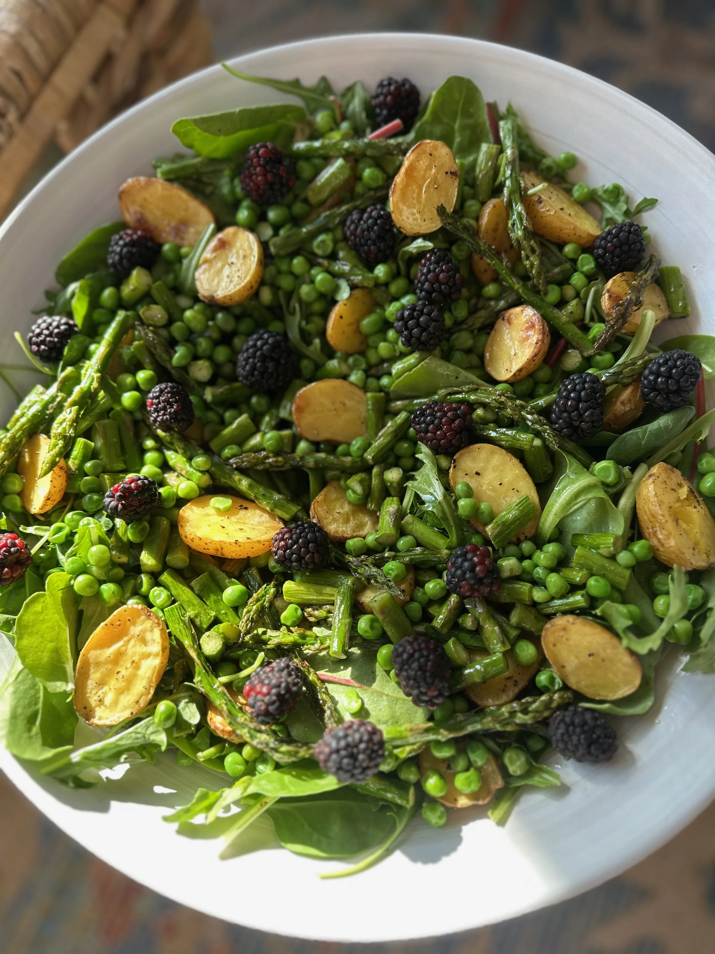 A white bowl filled with a salad containing green peas, roasted potato slices, asparagus, blackberries, and fresh greens.