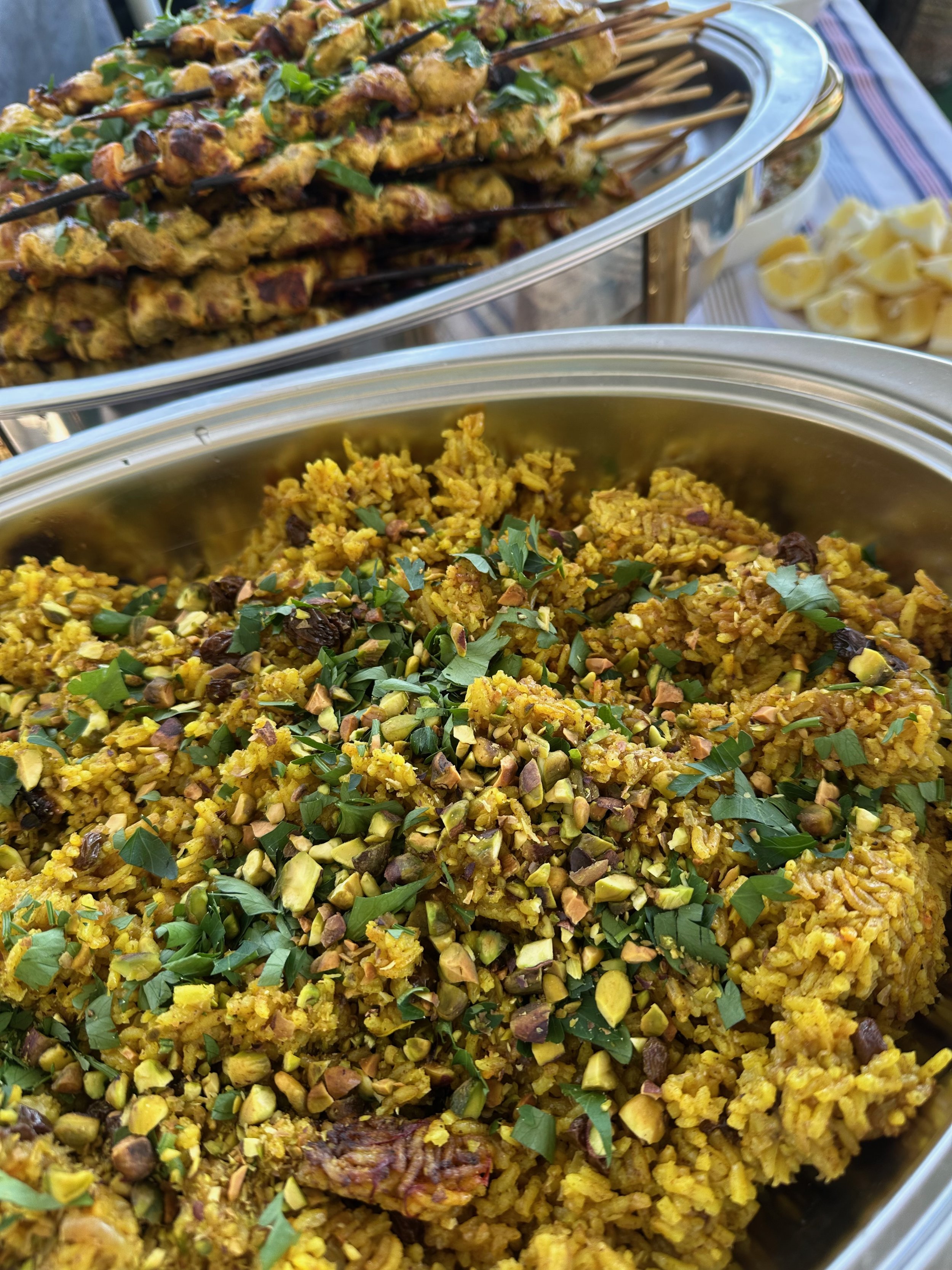 Close-up of a dish of yellow rice with chopped pistachios and herbs, and skewers of grilled meat garnished with herbs in the background.