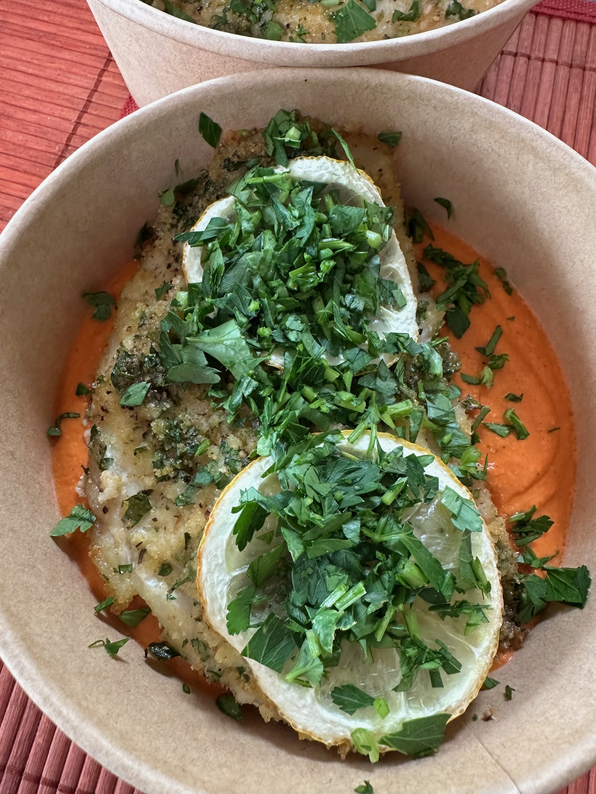 Baked fish fillet topped with chopped herbs and sliced lemon, served on a bed of orange sauce in a brown paper bowl.