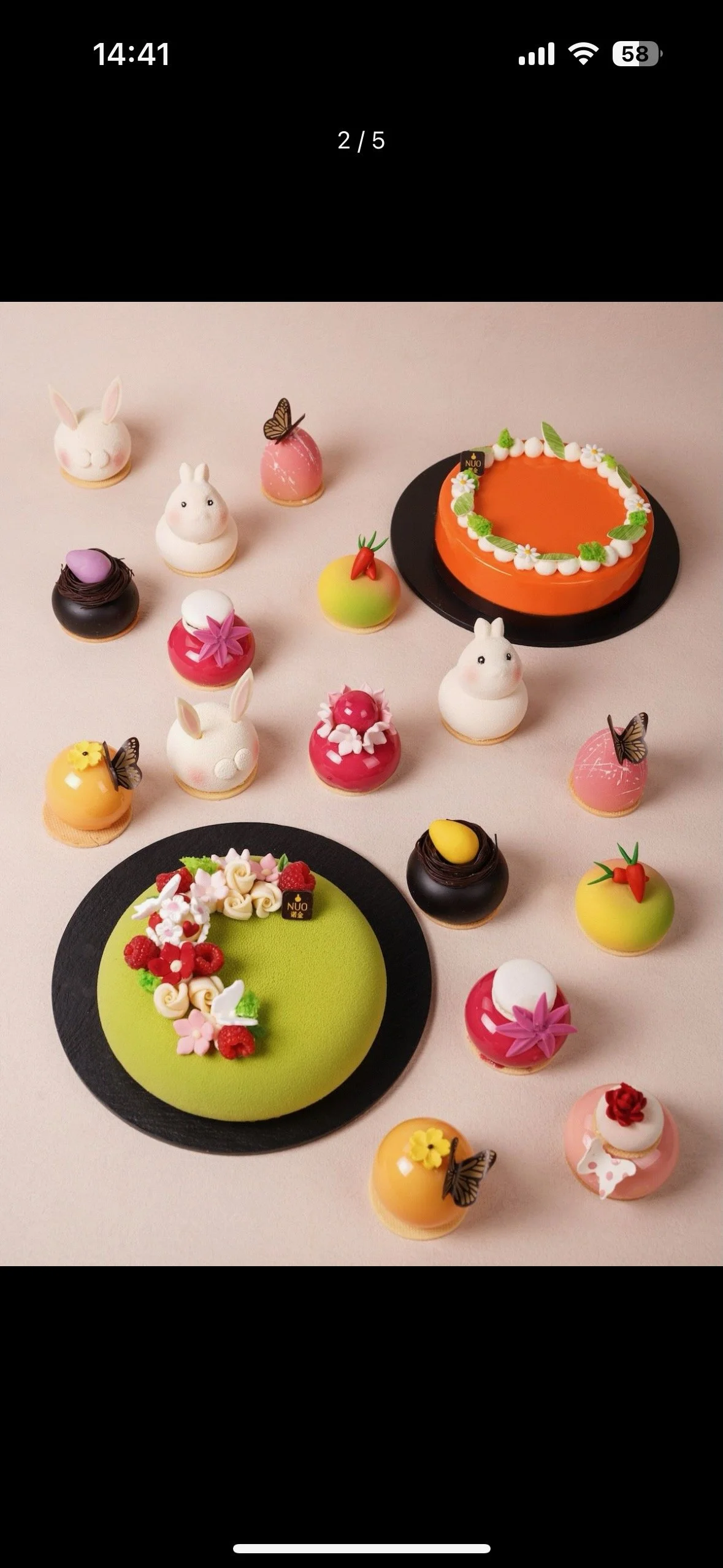 Assorted decorated mini cakes and pastries shaped like animals and fruits, with some featuring butterflies, on a light pink surface.