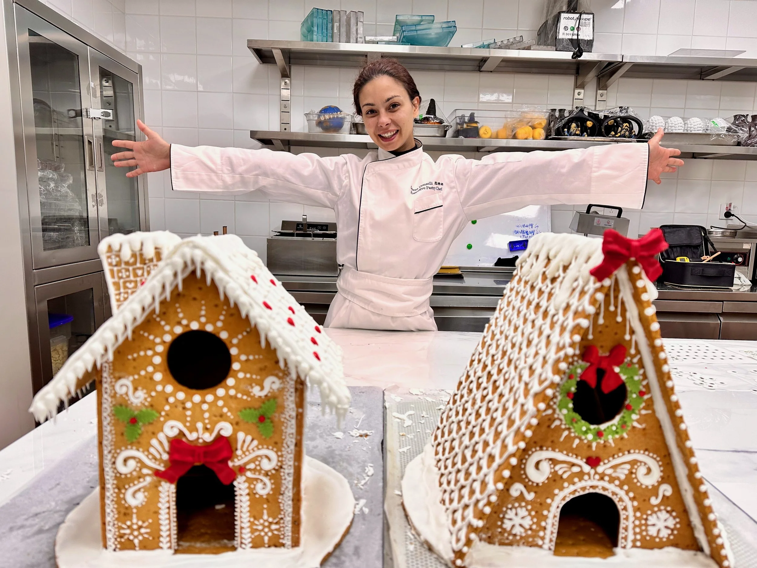 A woman in a white chef's coat stands in a kitchen with her arms outstretched, smiling behind two decorated gingerbread houses on a table. The gingerbread houses are adorned with white icing and red and green decorations, one with a red bow and the o